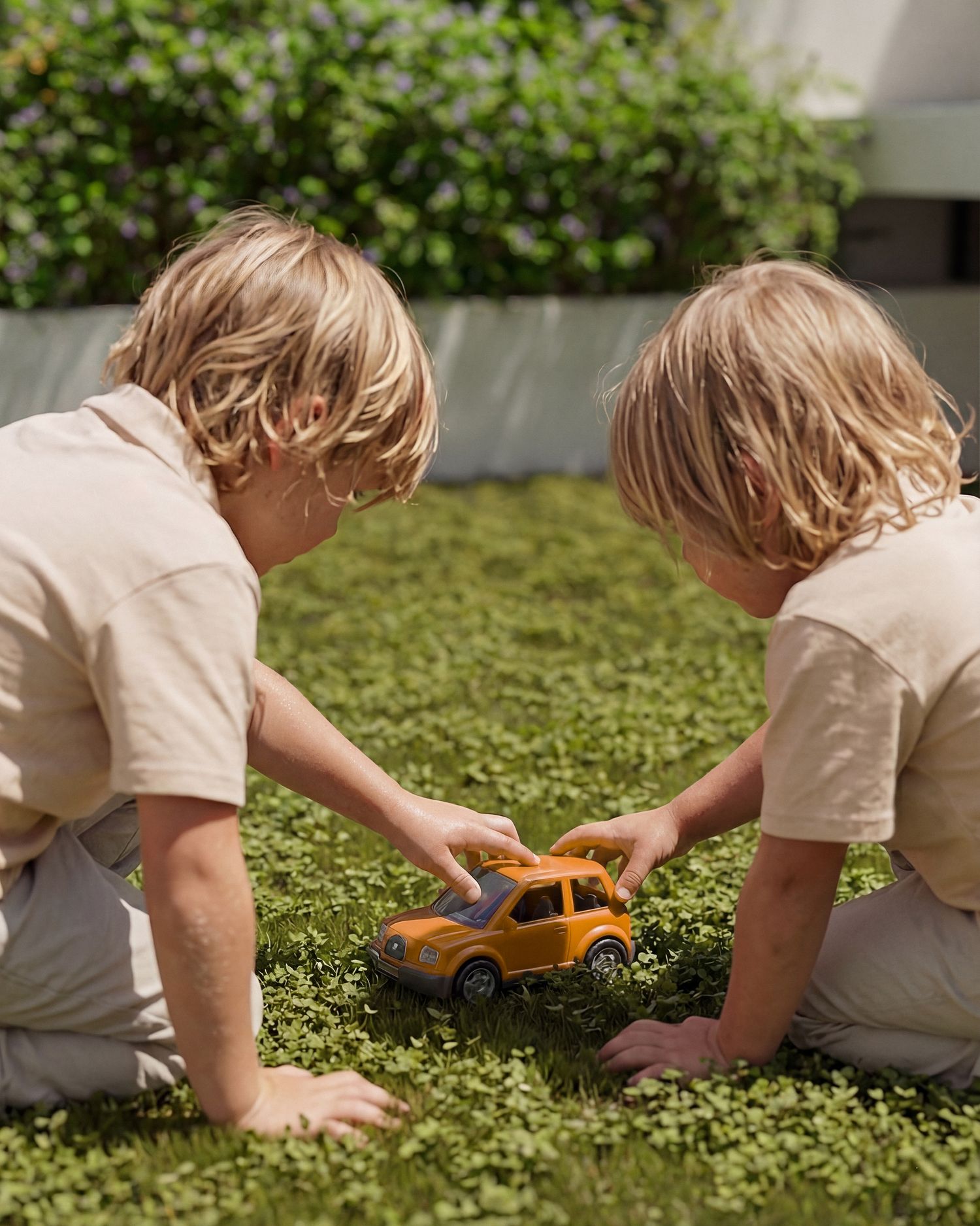 Two blonde twin boys in matching beige outfits playing with an orange toy car on a lush green lawn.