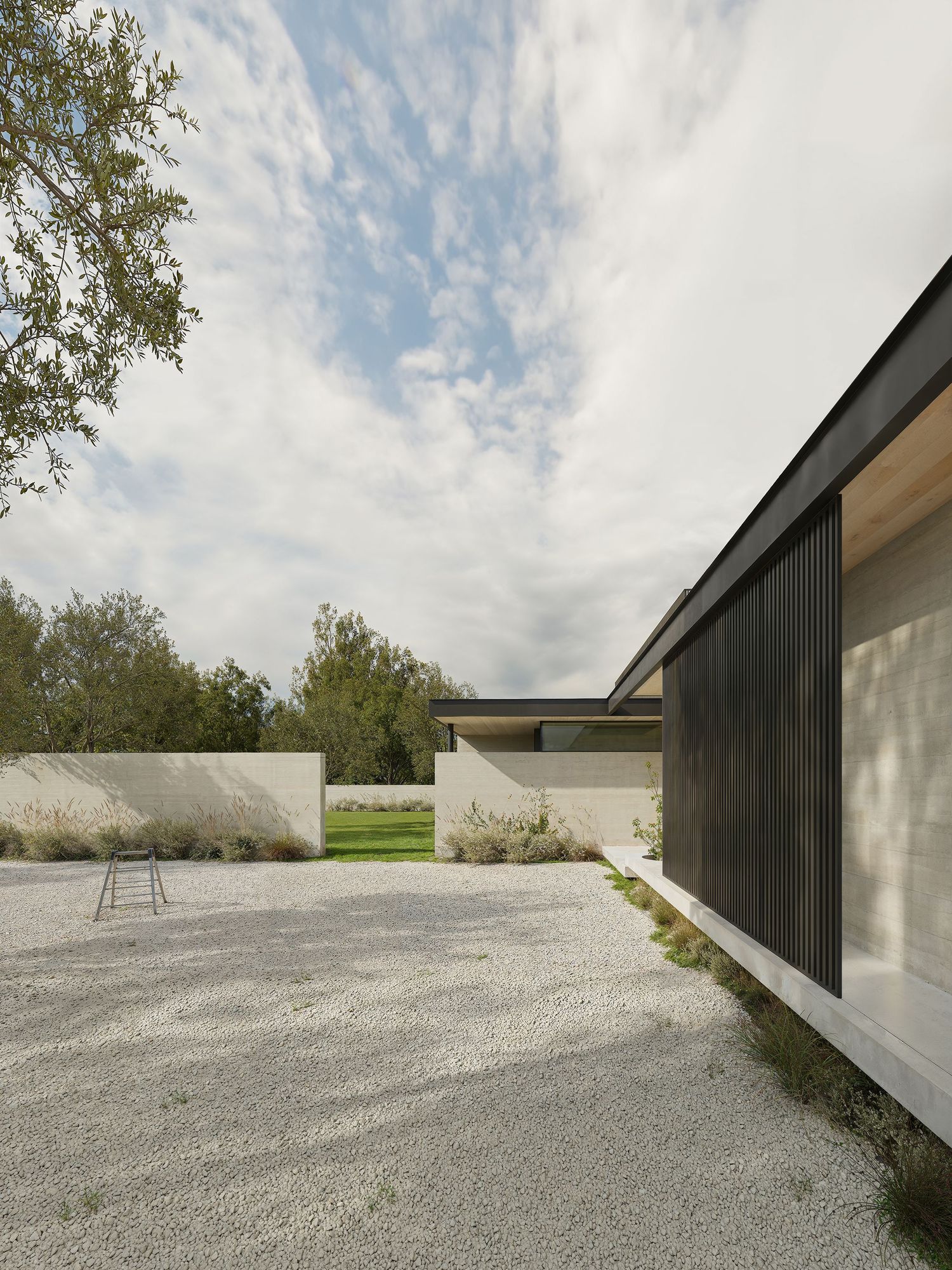 Modern minimalist house with black vertical siding and concrete courtyard set against cloudy sky and natural landscape.