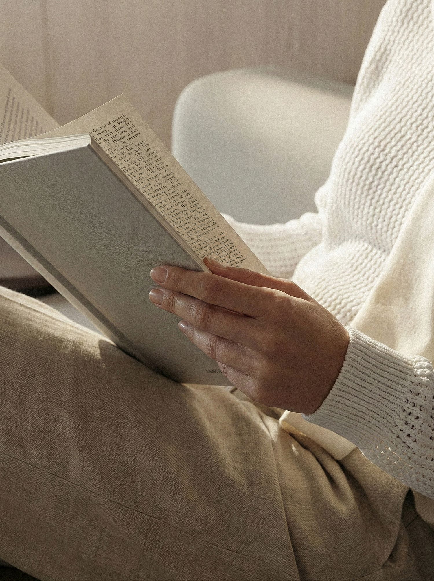 Someone in a white sweater holds an open book while sitting on a gray couch, creating a cozy reading atmosphere.