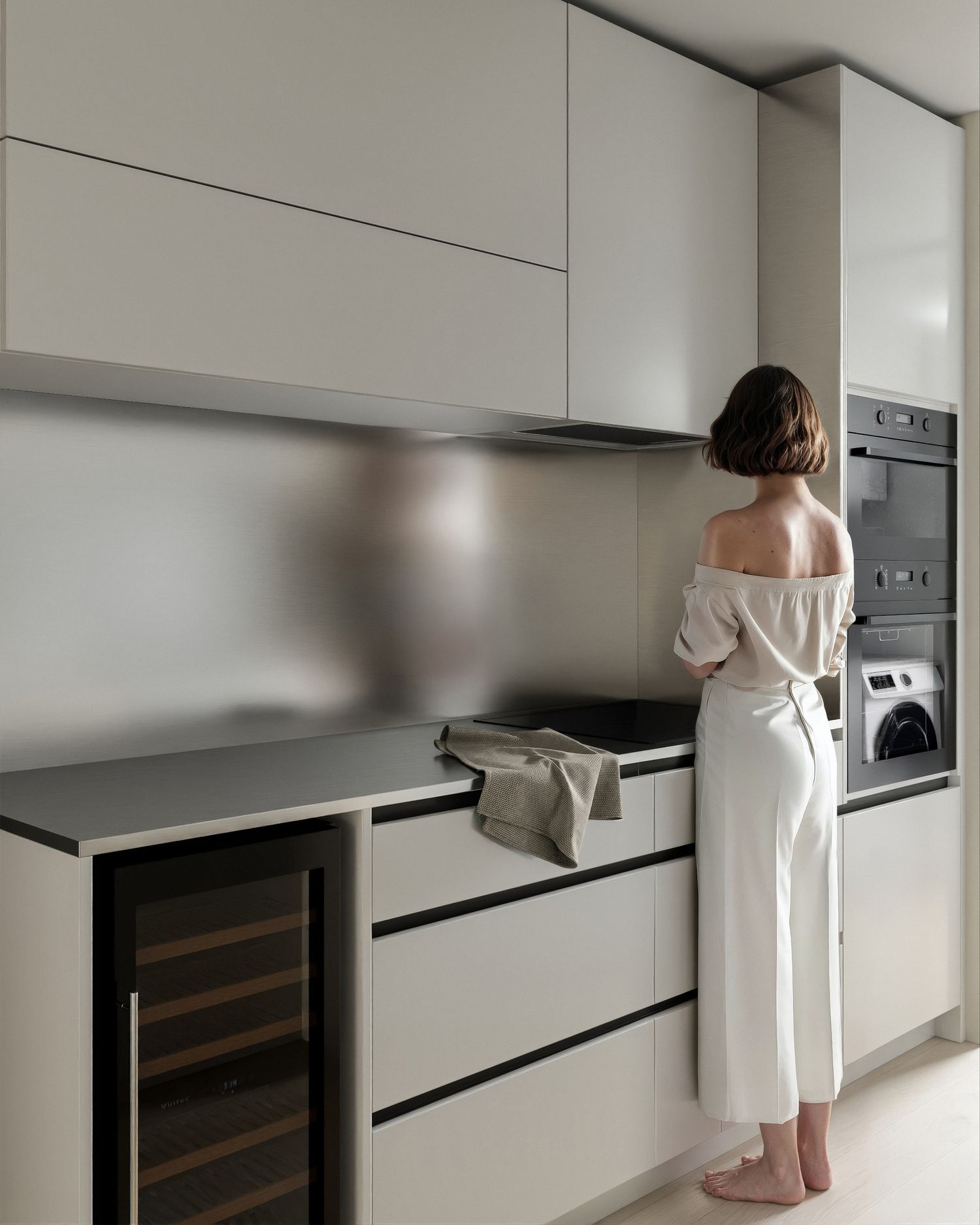Woman in white off-shoulder outfit standing at a modern minimalist kitchen with grey cabinetry and built-in appliances.