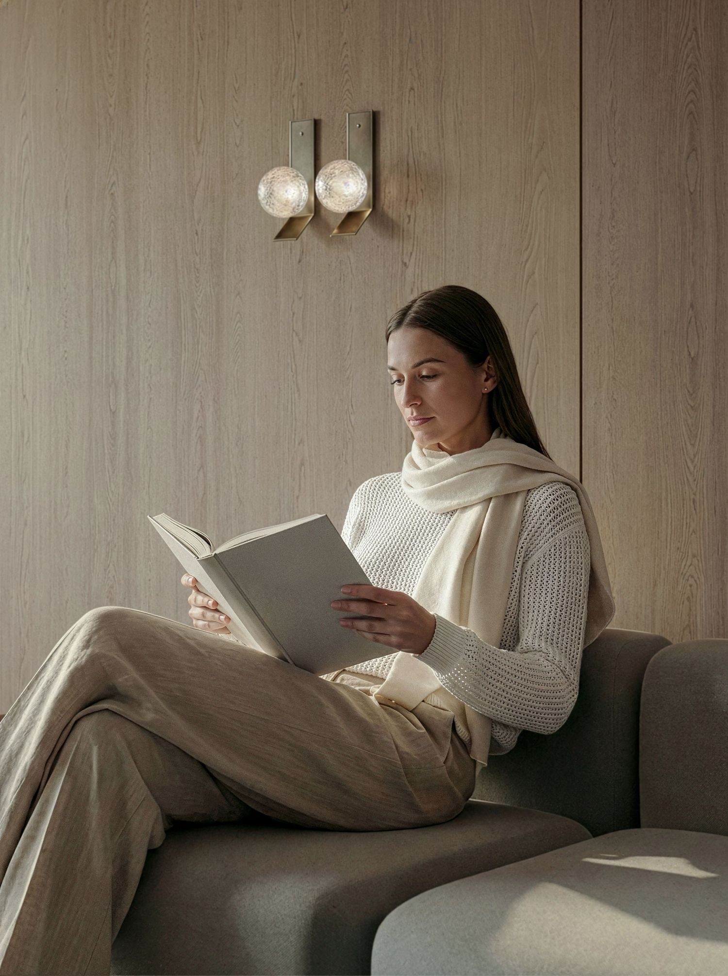 A person in a white sweater relaxes on a beige couch while reading a book in soft natural lighting.