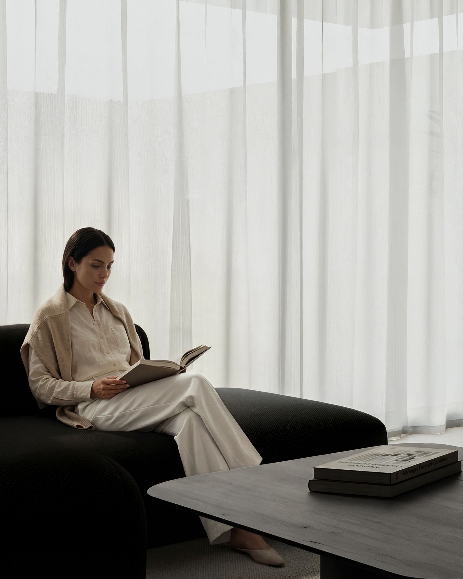 Woman in cream outfit reading a book on a dark sofa by sheer white curtains in a minimalist living room.