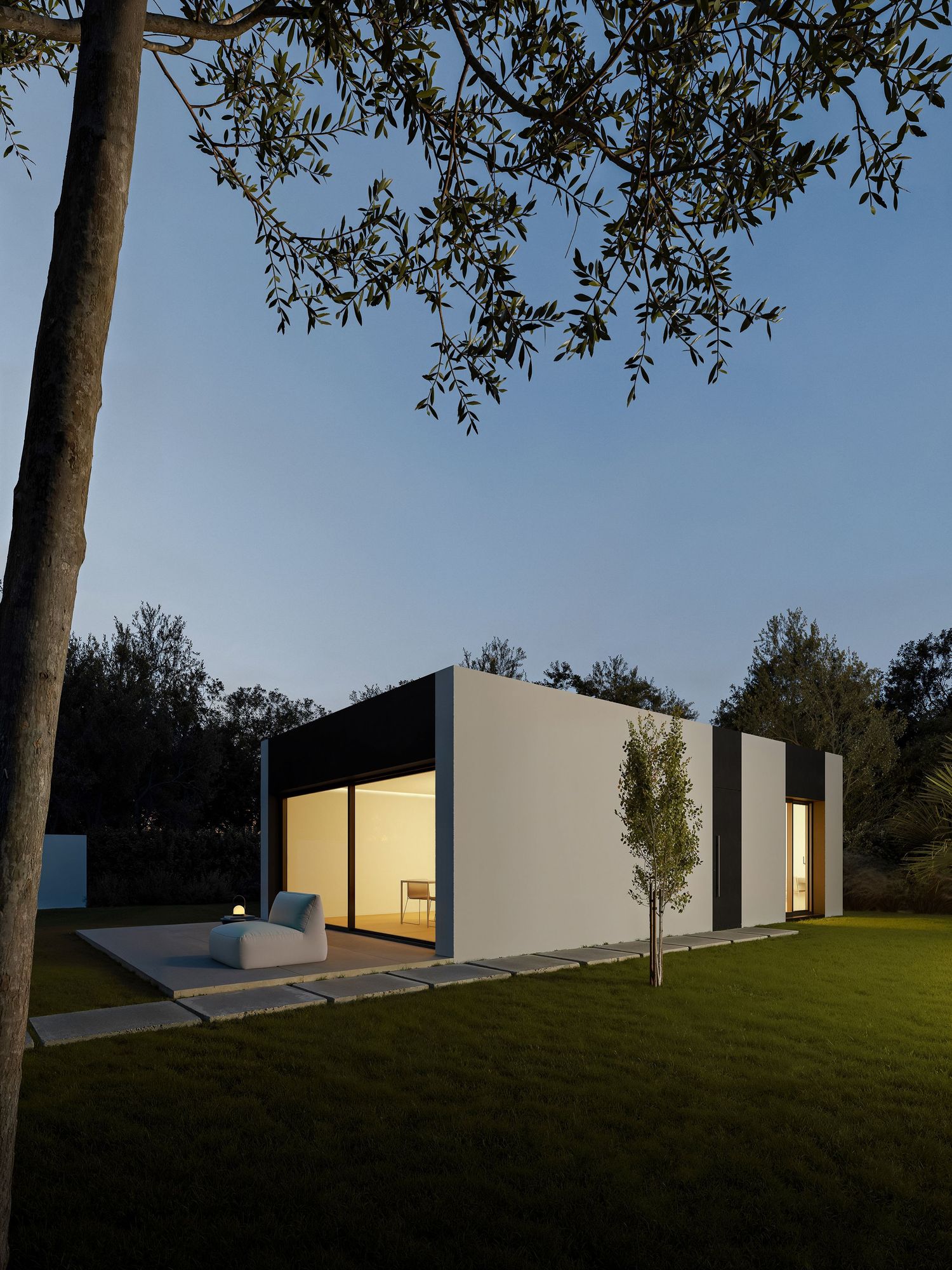 Modern minimalist white house with large windows and illuminated interior viewed at dusk under an olive tree.
