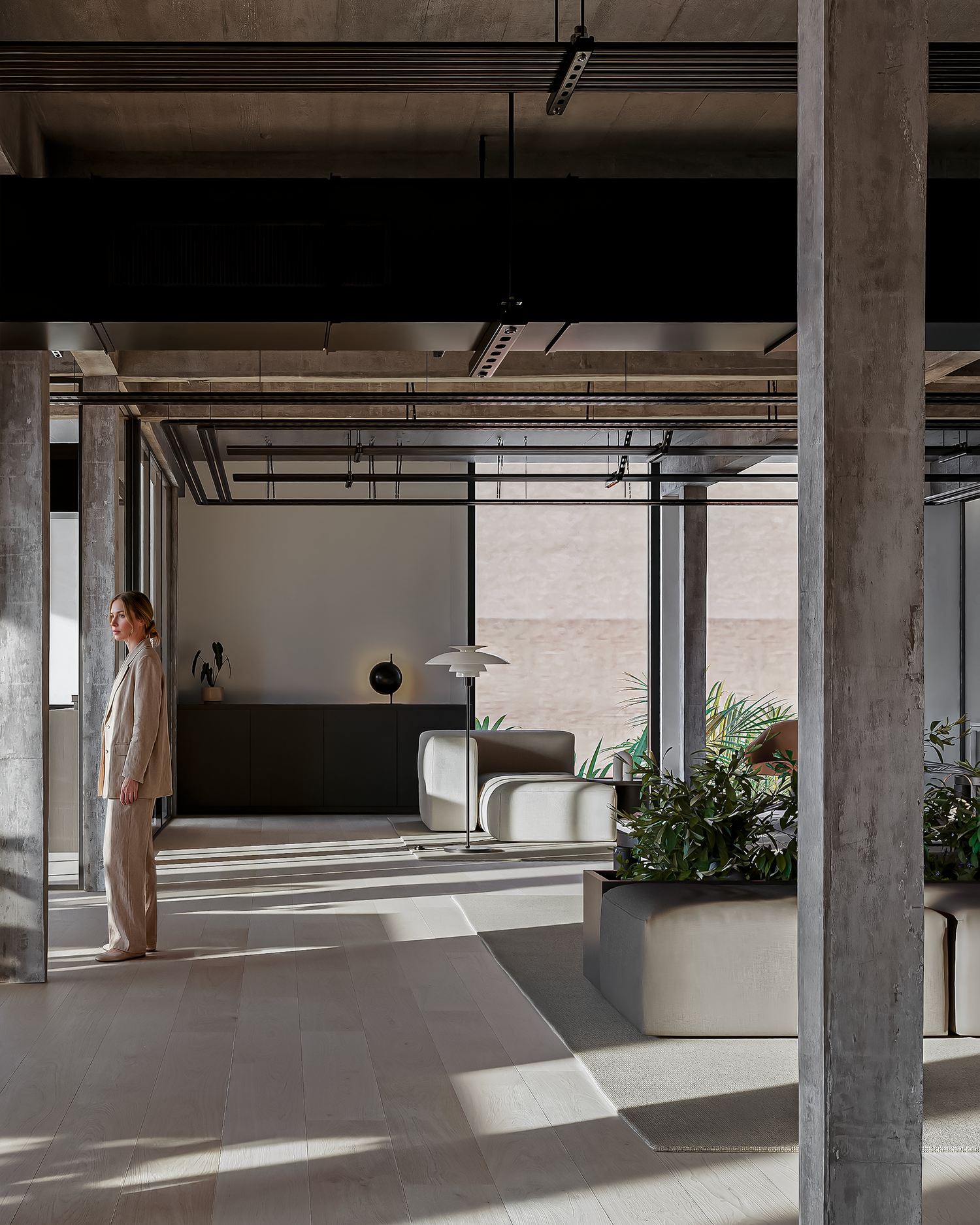 Modern minimalist office lobby with concrete columns, lush plants, and a woman standing near a dark reception desk.