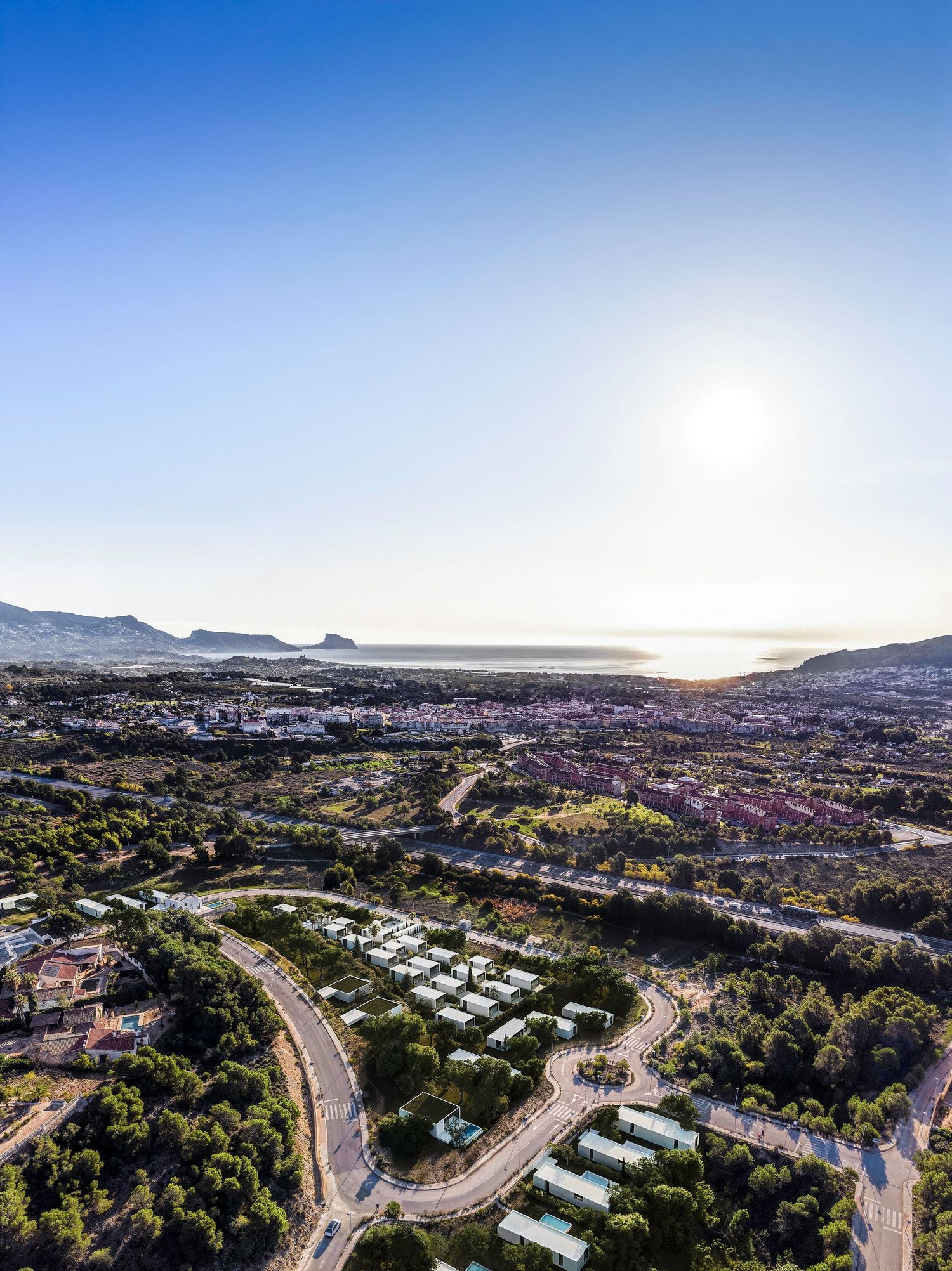 Aerial view of a coastal residential community with winding roads and houses nestled in hills overlooking the ocean at sunset.