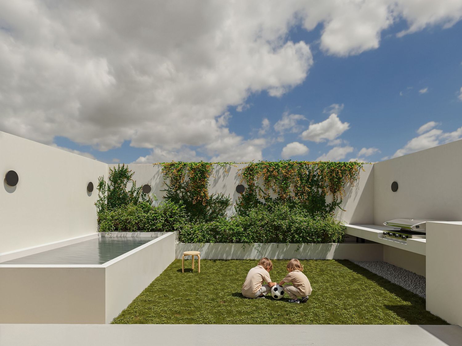 Modern rooftop garden with green wall, pool, lawn, and two people relaxing under a cloudy blue sky.