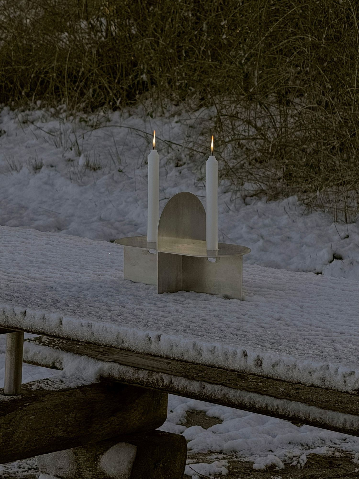 Two white candles stand on a small platform over a snowy bridge in a winter landscape.