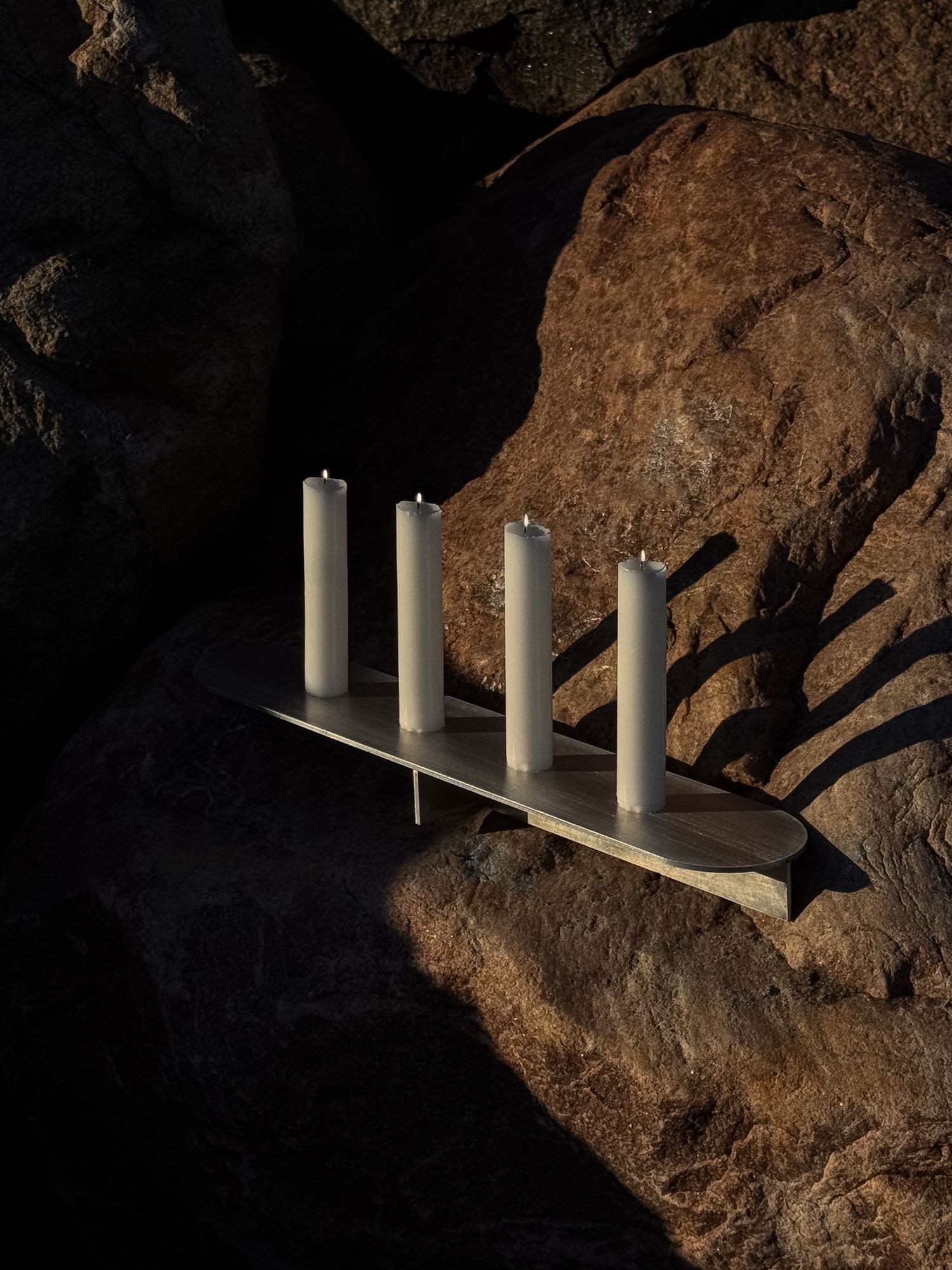 Four white pillar candles arranged on a minimalist metal shelf against a textured rock surface in dramatic lighting.