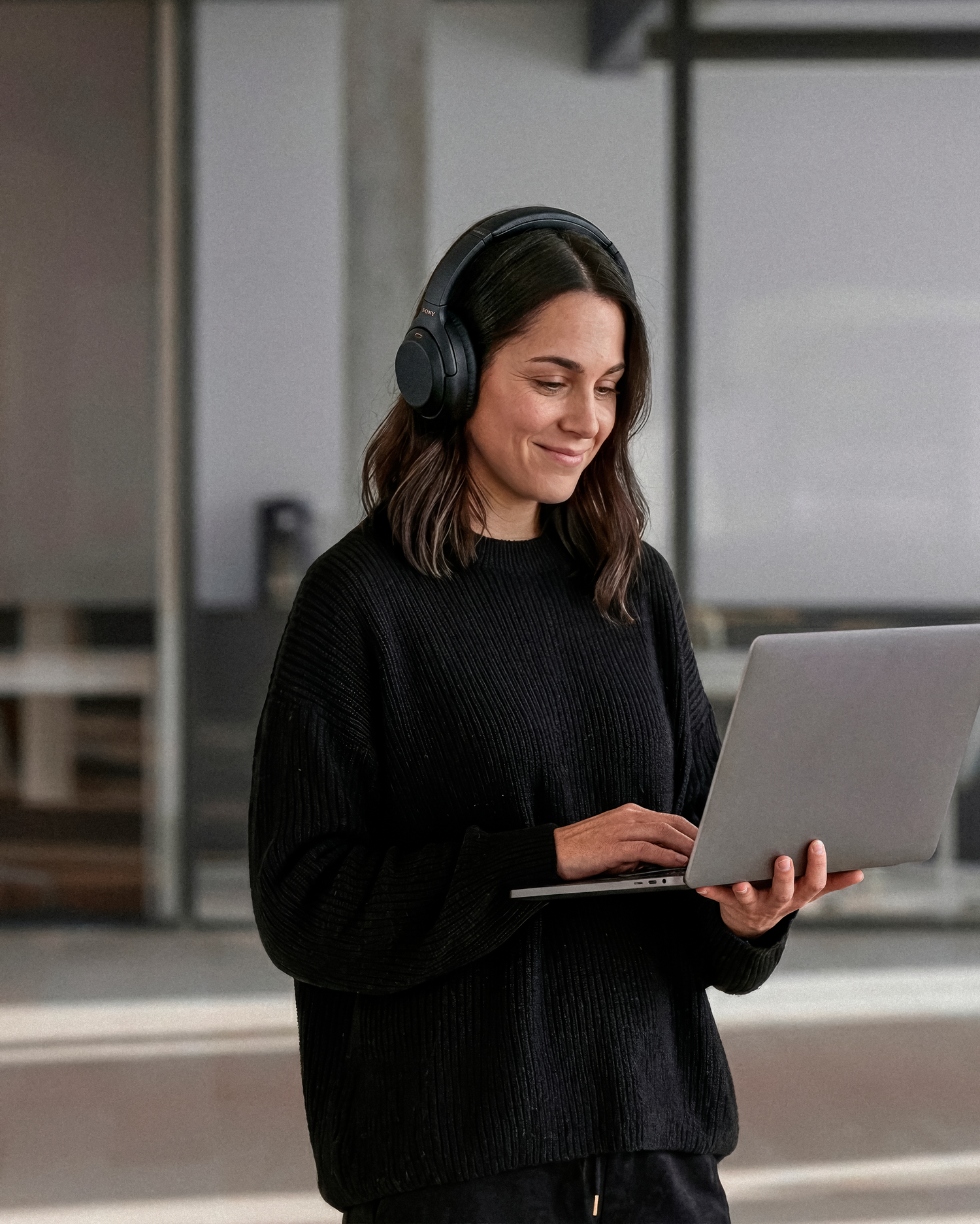 Woman in black sweater wearing headphones smiles while using a laptop in a modern office setting.