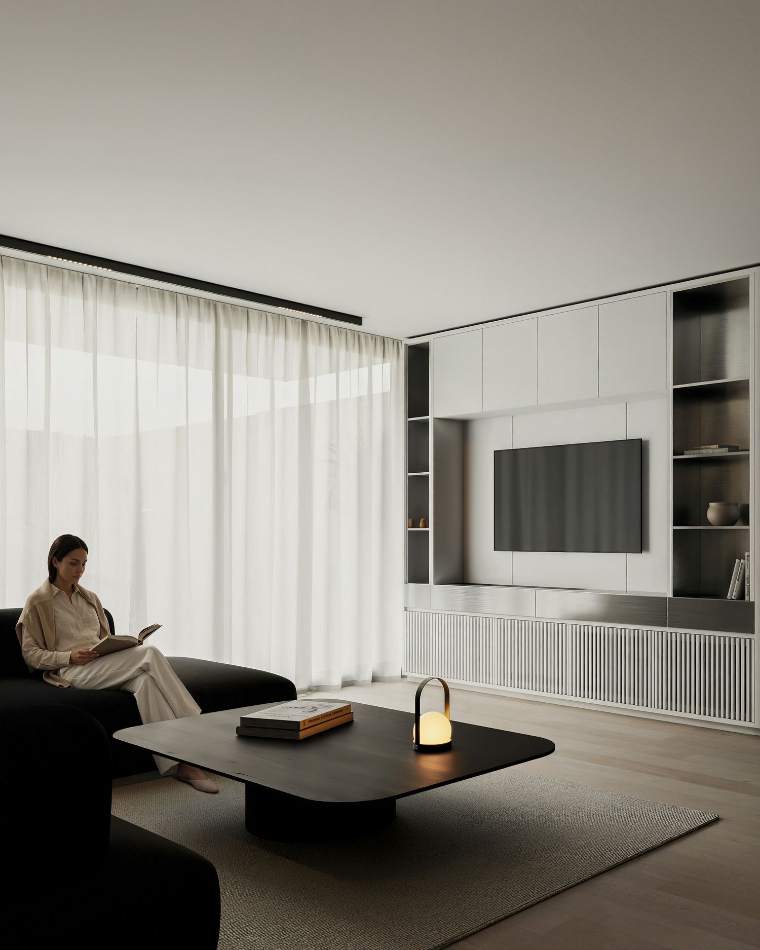 Minimalist living room with built-in shelving, wall-mounted TV, sheer curtains, and a woman reading on a black sofa.