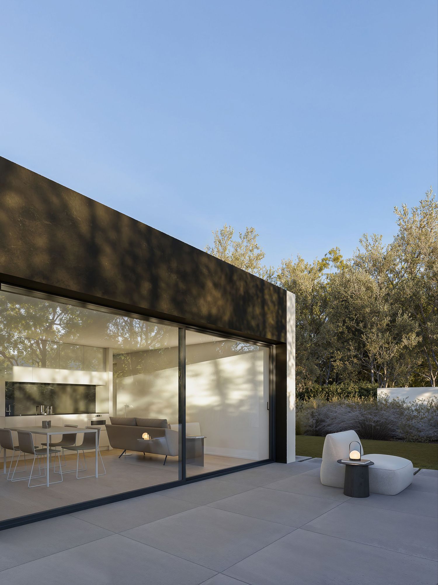 Modern outdoor living space with glass sliding doors opening to a minimalist patio featuring contemporary furniture under clear skies.