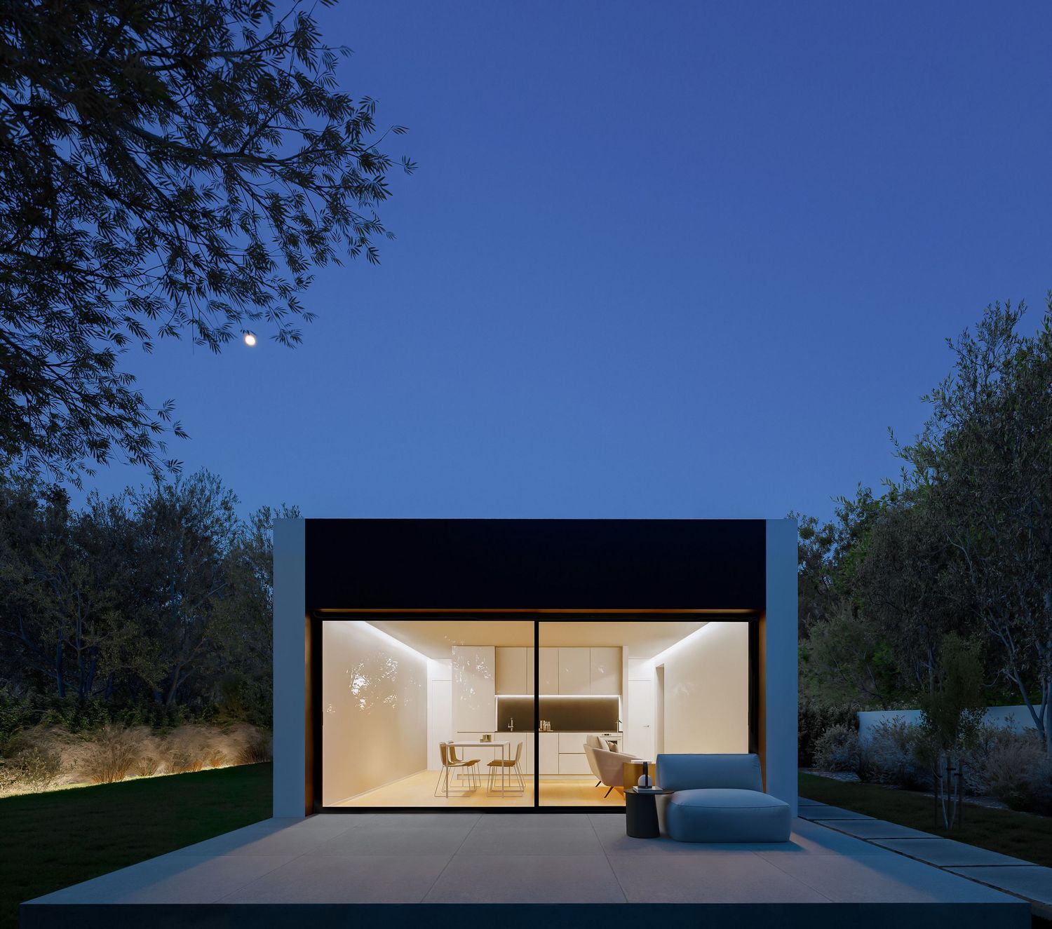 Modern minimalist home illuminated at dusk with large glass windows showing interior lighting against darkening sky.