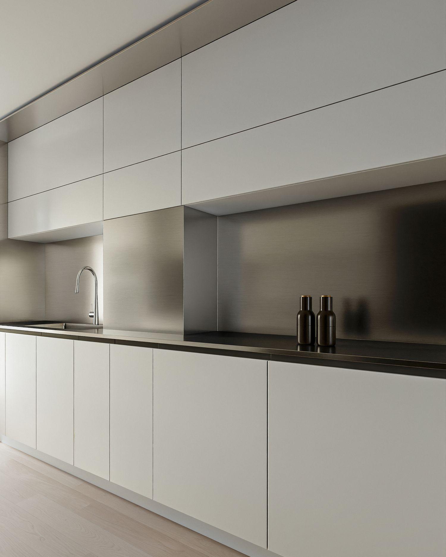 Minimalist kitchen with white handleless cabinets, dark countertop, stainless steel backsplash, and two black bottles.