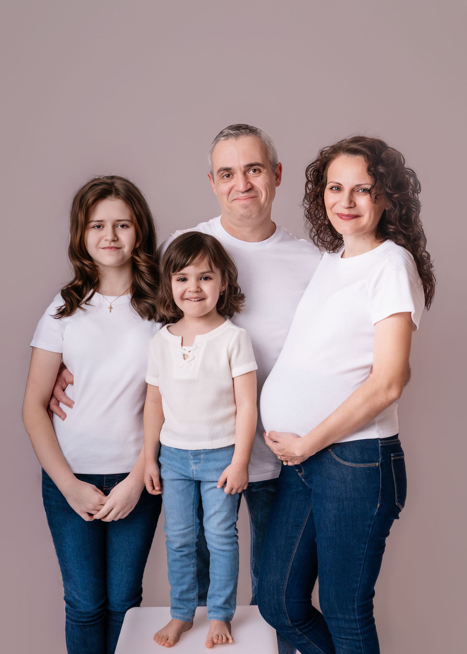 Family portrait with pregnant mother, father, and two daughters in white tops and jeans on a neutral background.