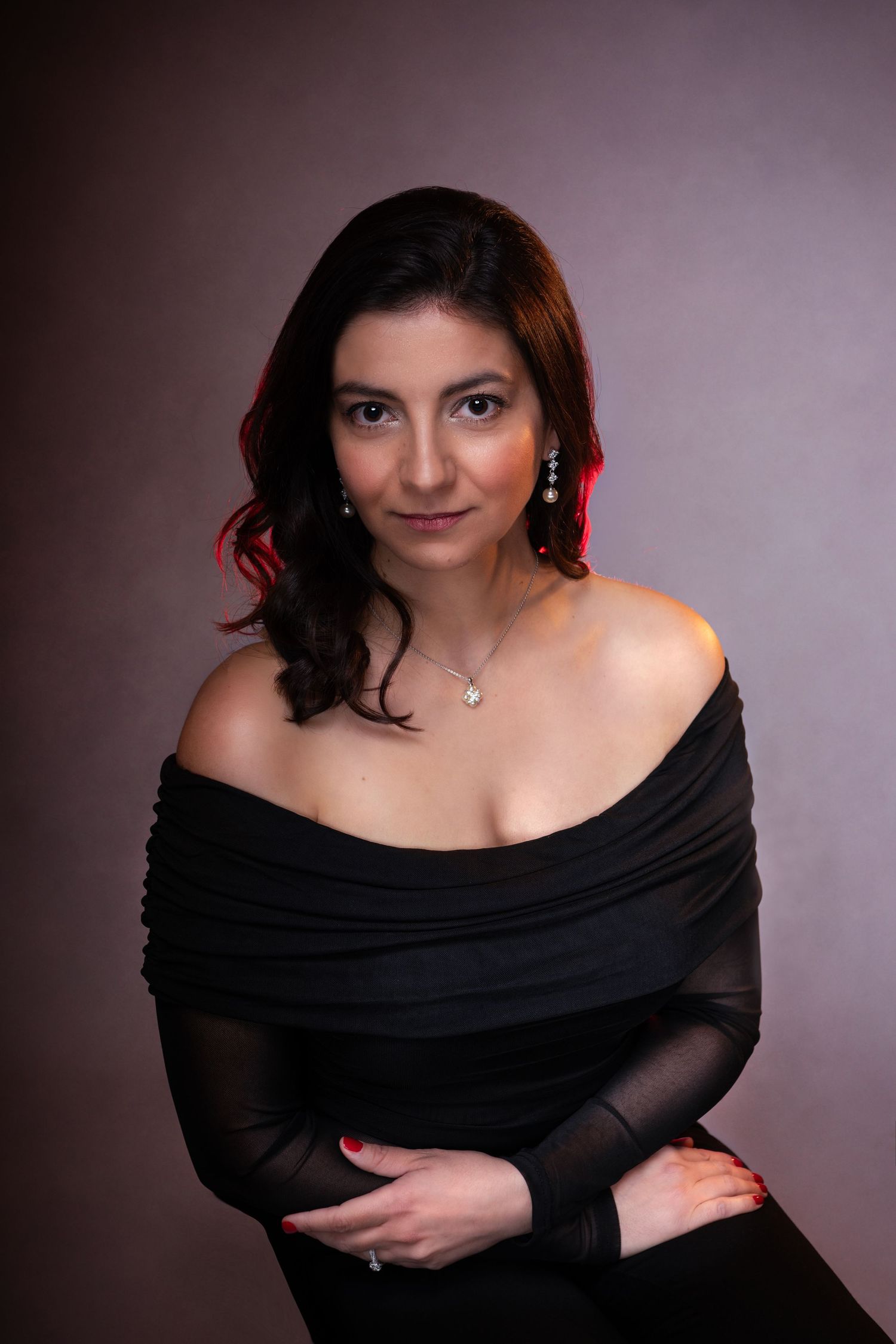 Woman in black off-shoulder top with silver necklace and earrings poses elegantly against a dark studio background.