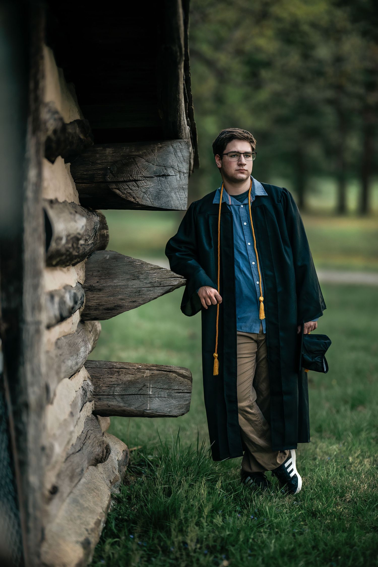Young man in graduation gown and gold honor cord poses by rustic log cabin wall outdoors.