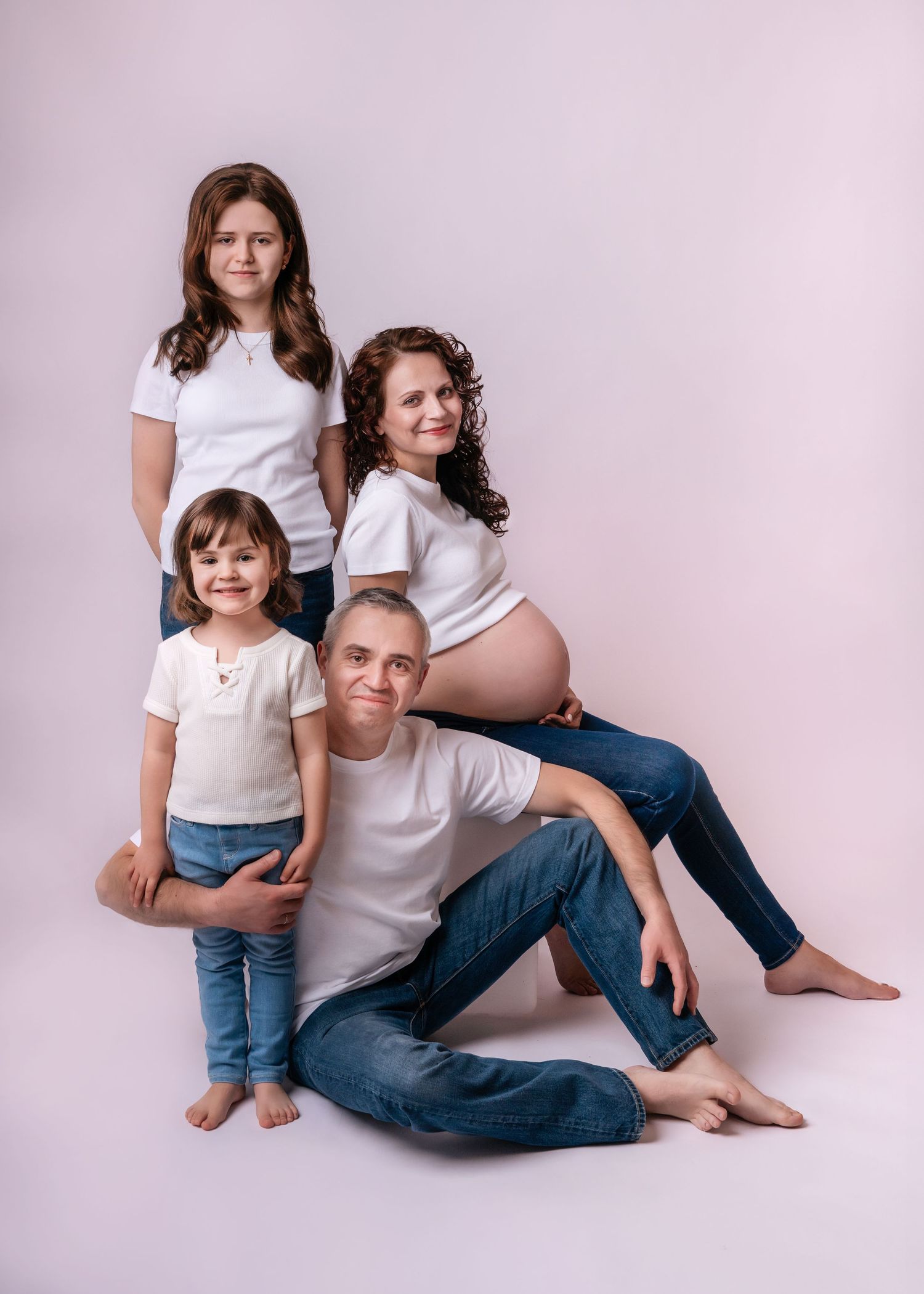 Family maternity photo with pregnant mother, father, and two daughters in matching white tops and jeans on light background.
