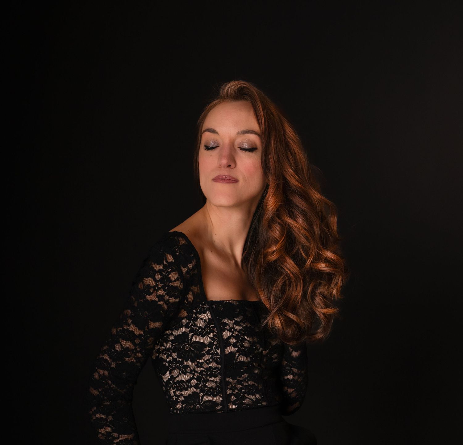 Woman in black lace dress with long auburn wavy hair poses gracefully against dark background.