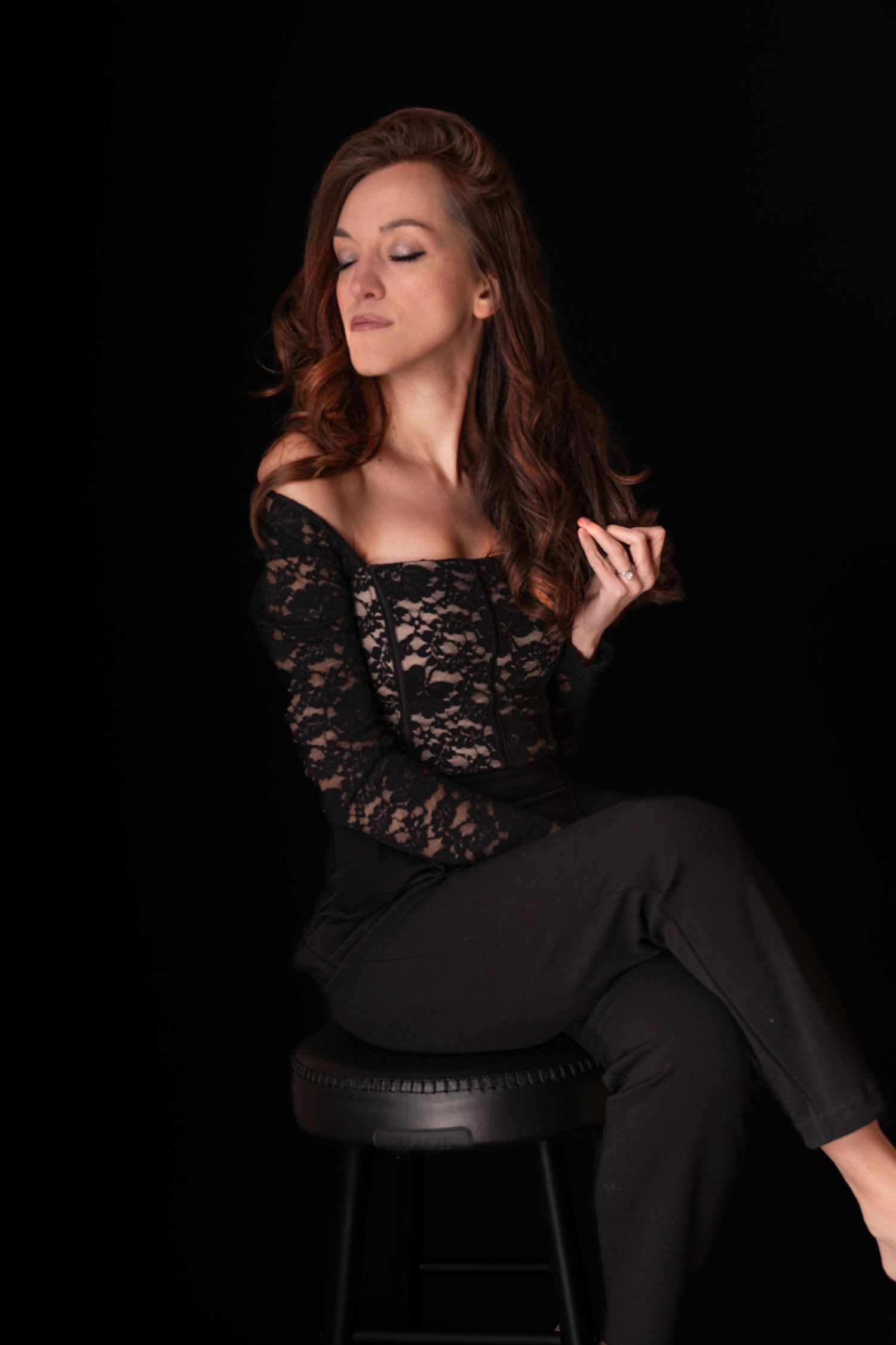 Woman in black lace off-shoulder top and pants sitting gracefully on a stool against a dark background.