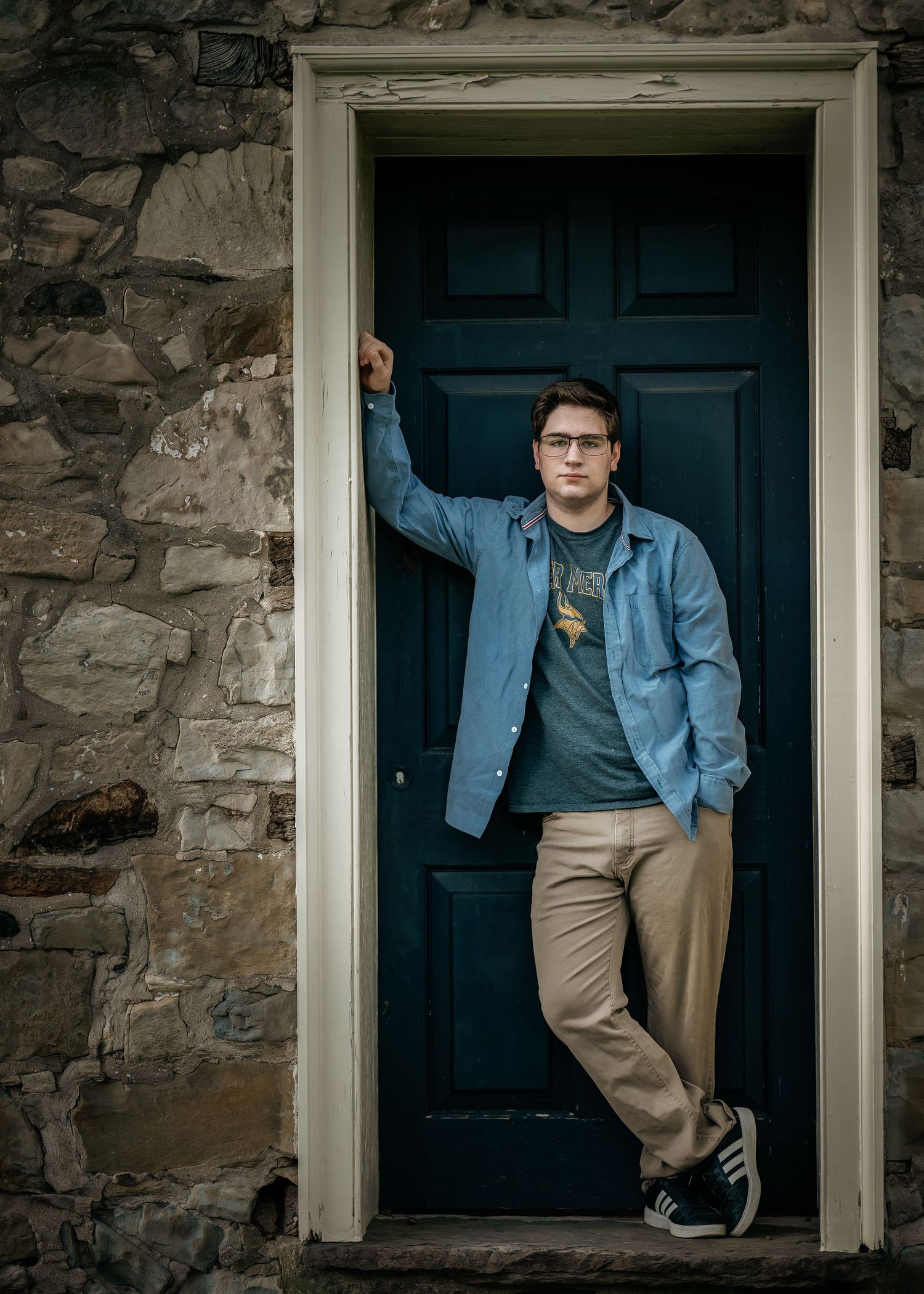 Young man in blue shirt and khakis leaning against a teal door in a stone building doorway.