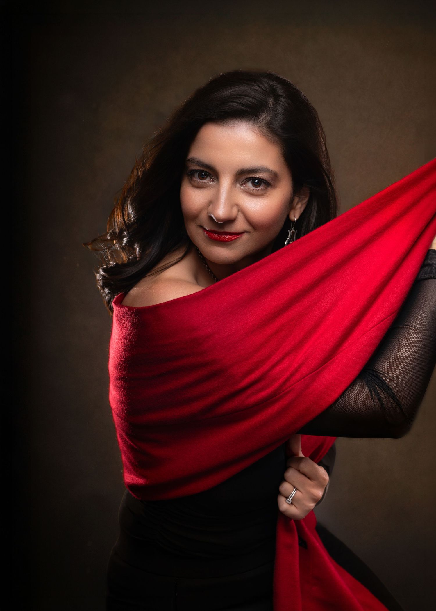 Woman in red draped fabric and black dress poses elegantly against a dark studio background during her glam portrait session in Wayne, PA..
