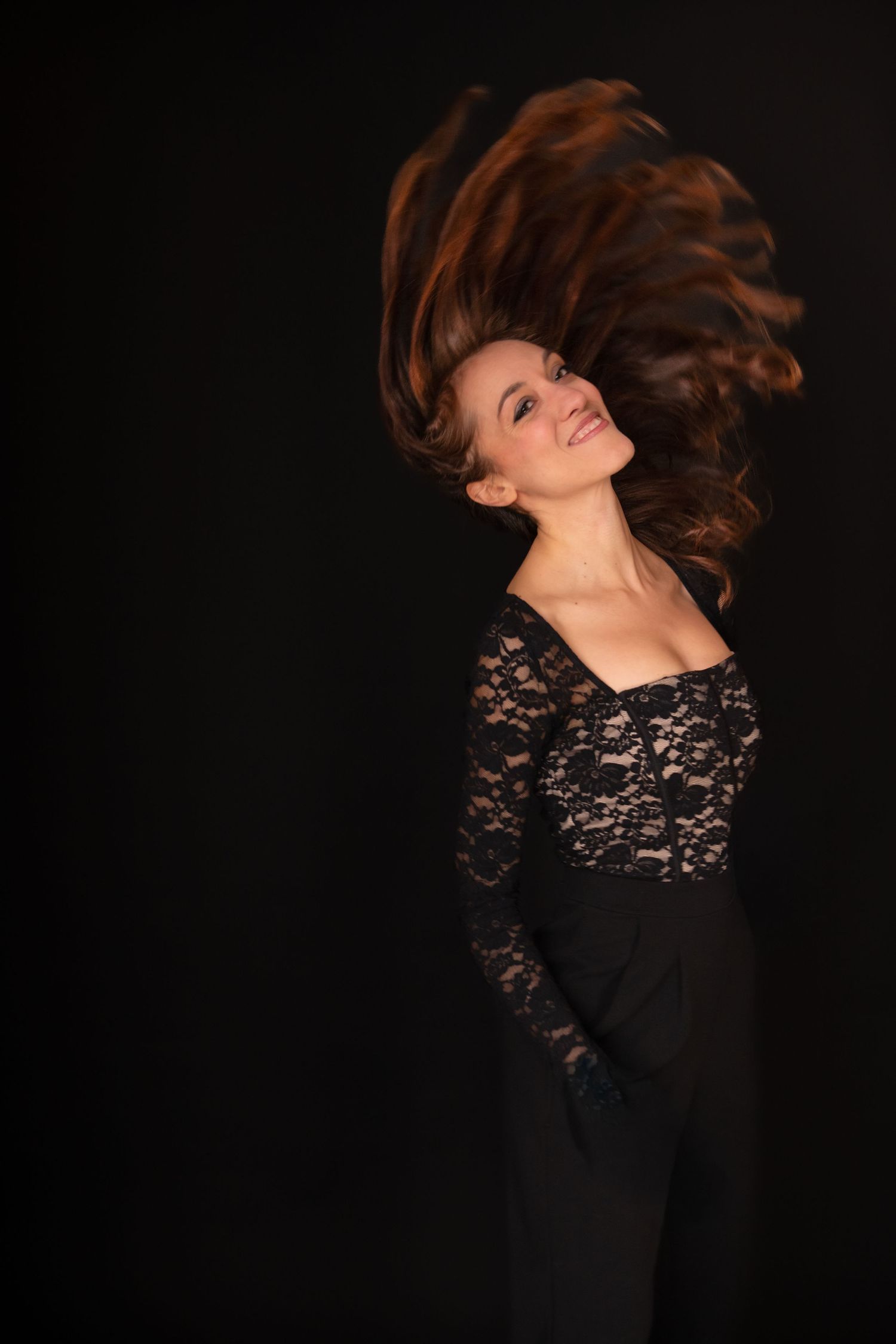 Woman in black lace jumpsuit tossing long auburn hair against dark background, smiling joyfully.