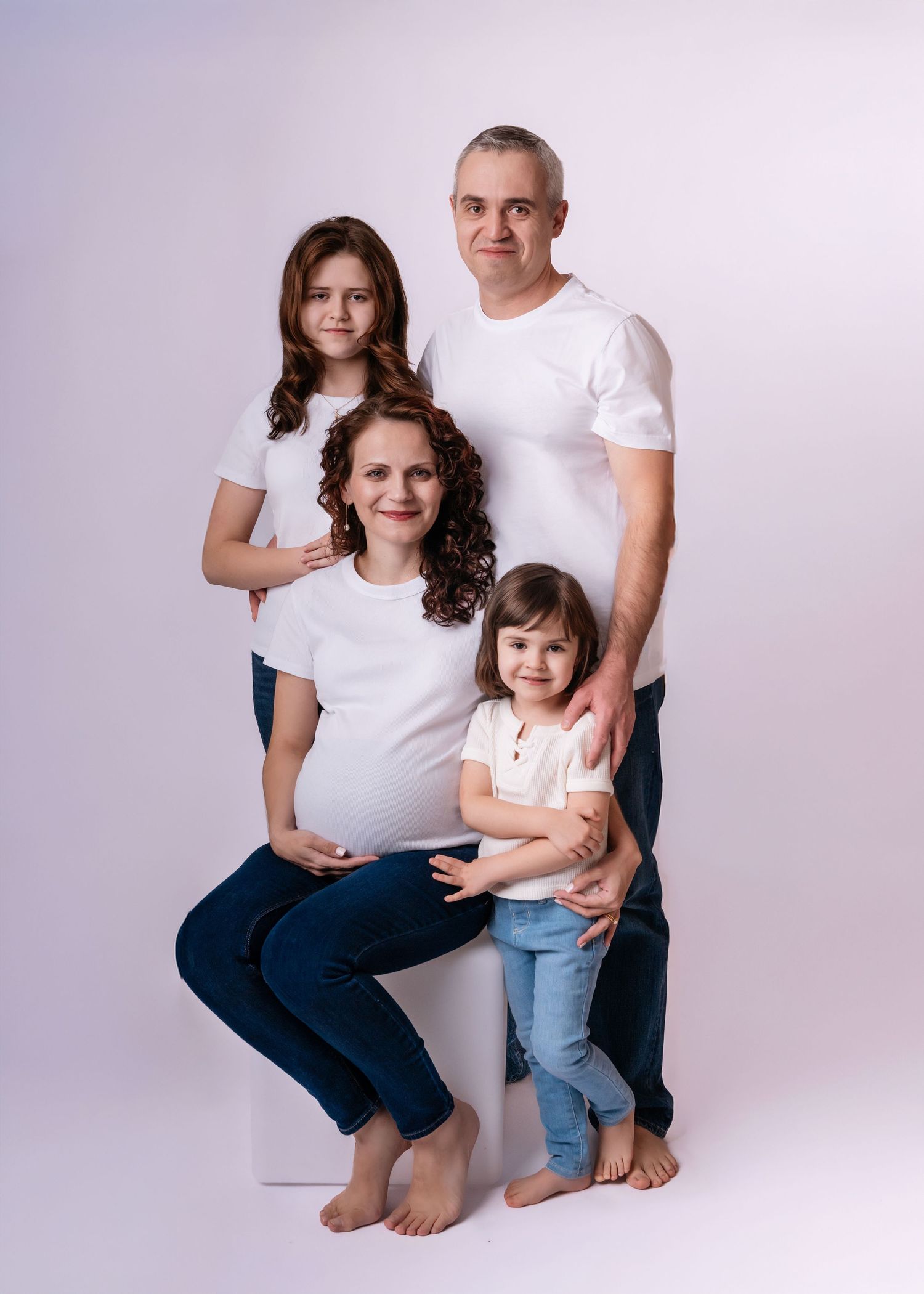 A pregnant woman in white shirt poses with her husband and two daughters in matching white tops and jeans for a family portrait.