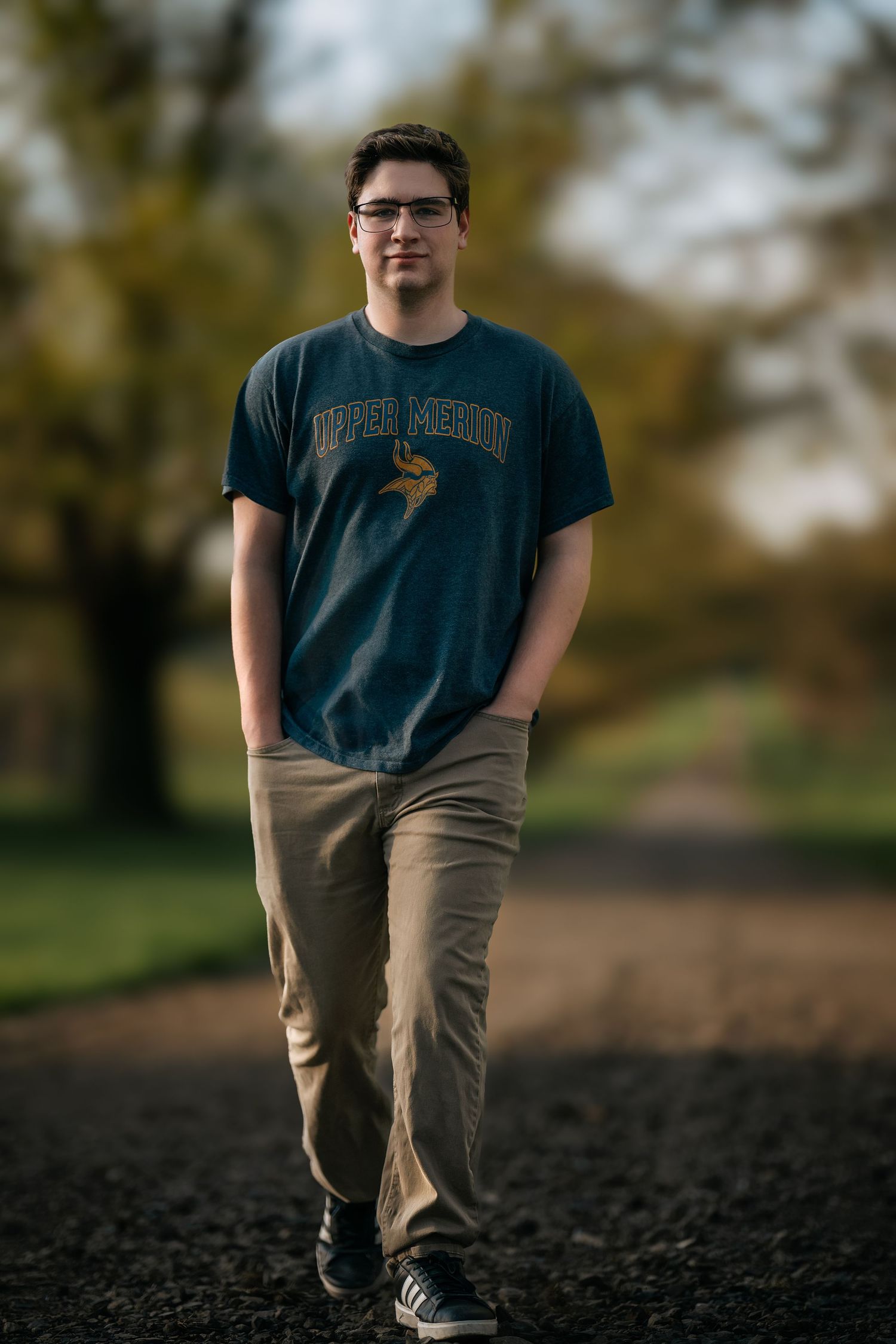 Young man in Upper Merion t-shirt and khakis walking on a gravel path in a park during autumn.