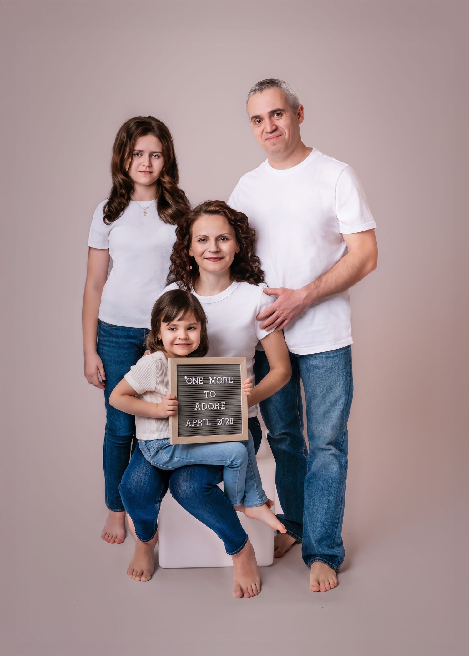 Family of four in white shirts holding a chalkboard sign announcing new baby in April 2028.