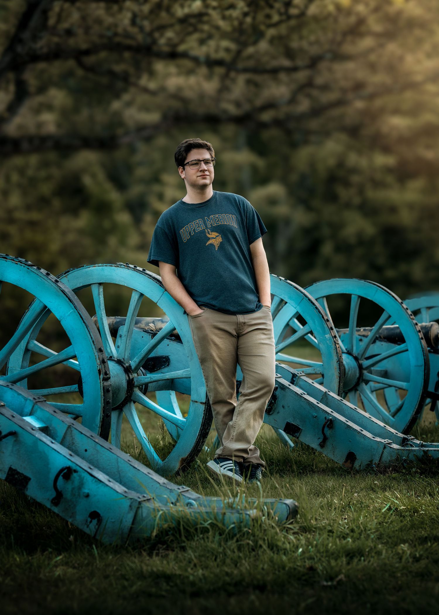 Young man in blue Vikings shirt stands among vintage blue cannons in a lush green outdoor setting.