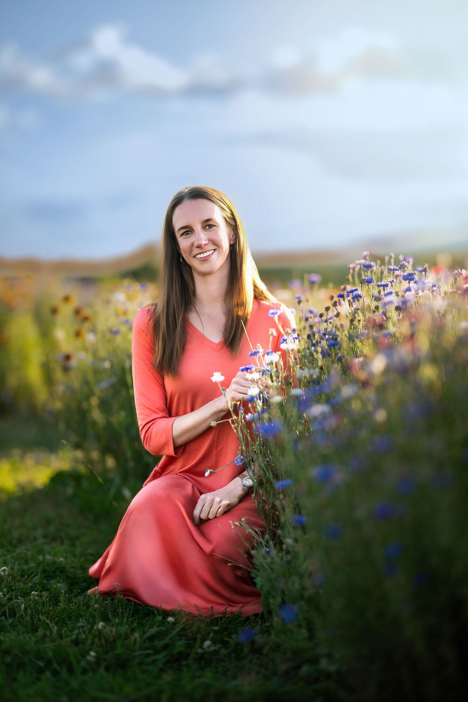 Smiling woman in red dress kneeling among colorful wildflowers in a sunlit field during an outdoor women's portrait session on the Main Line