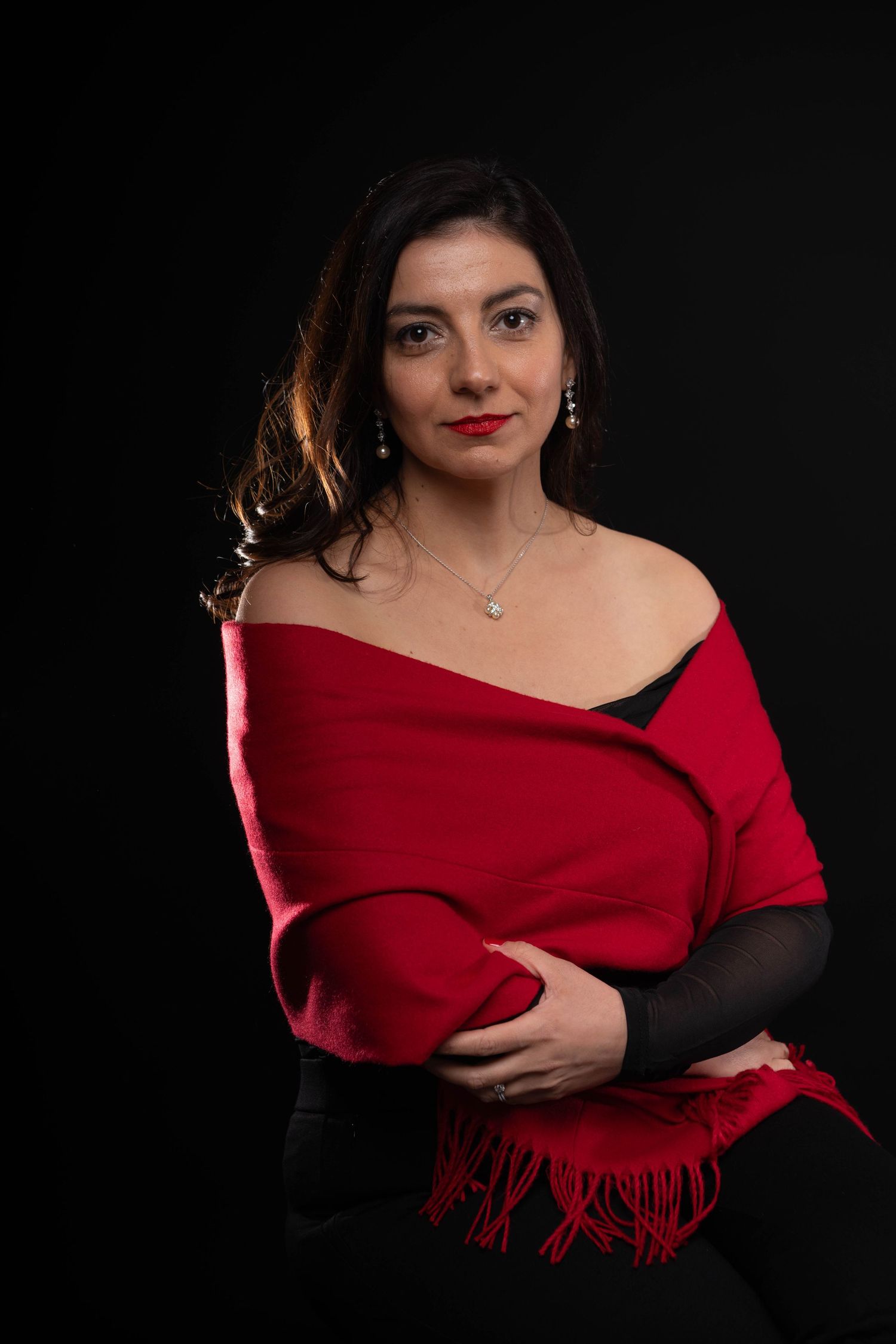Woman in red fringed shawl with red lips and diamond necklace poses against dark background.
