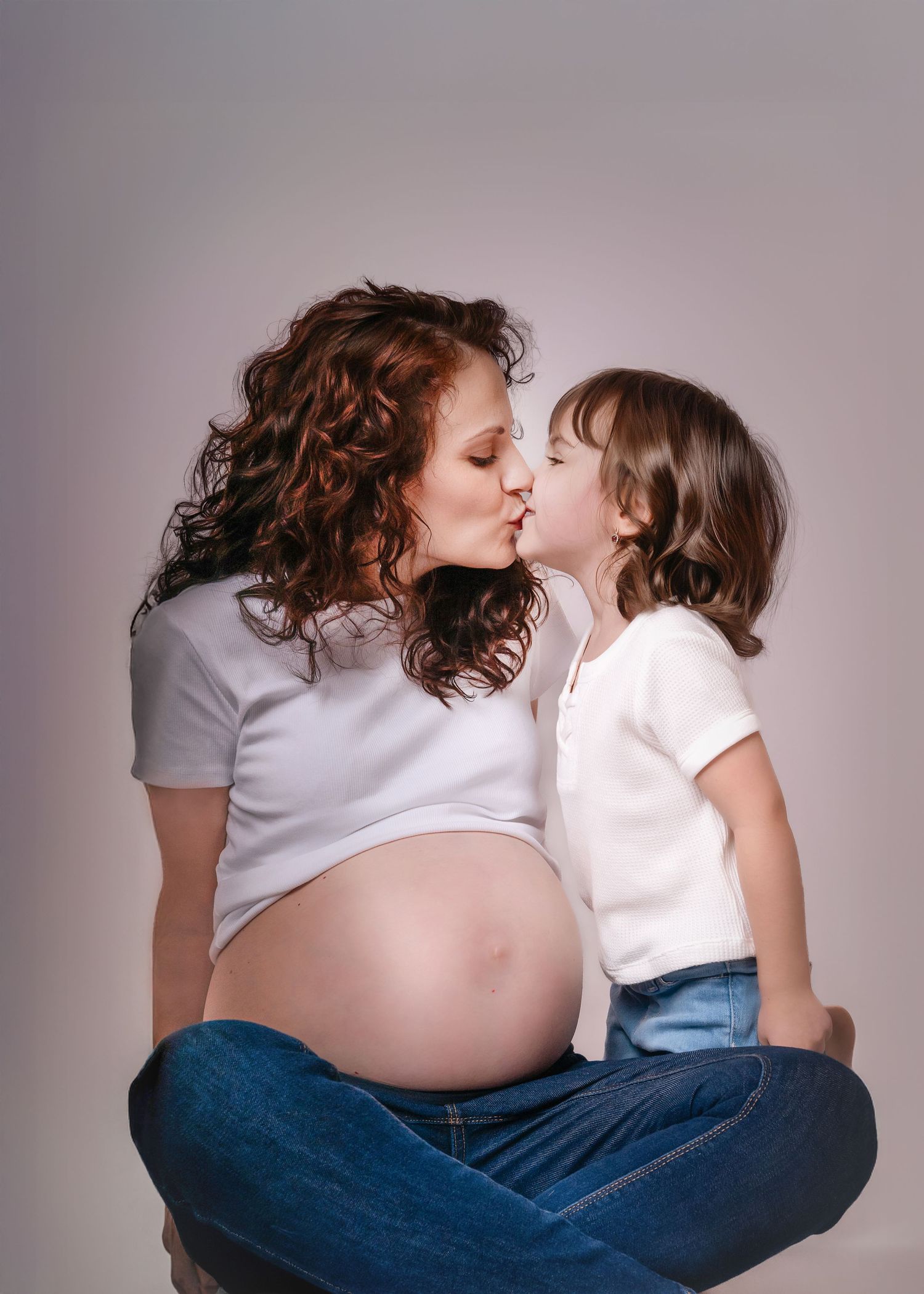 Pregnant mother with curly hair kissing young child during studio maternity photoshoot.