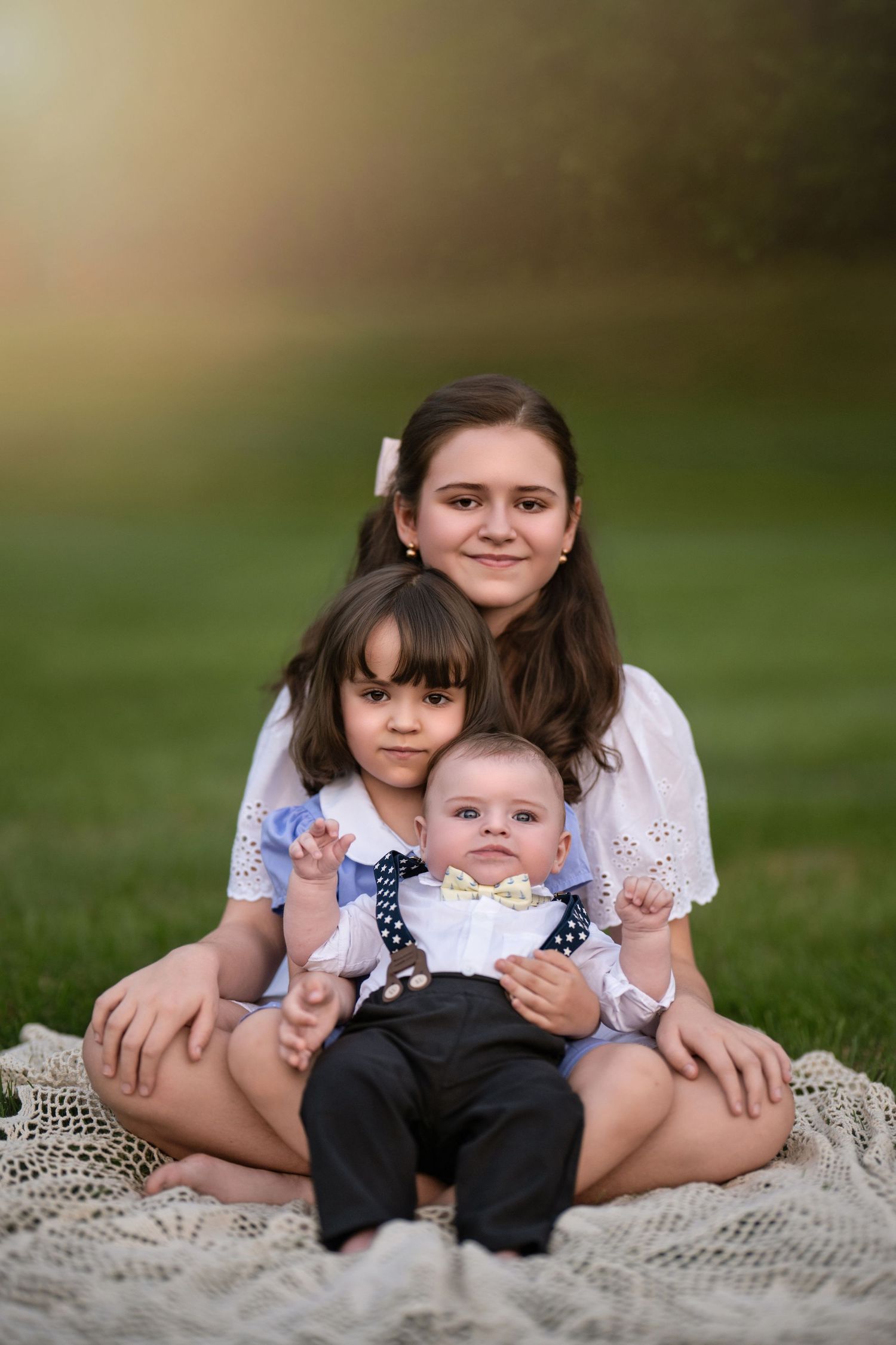 Three siblings pose outdoors on a blanket, two girls and a baby boy in formal attire, with a sunny green background near the Main Line, PA.