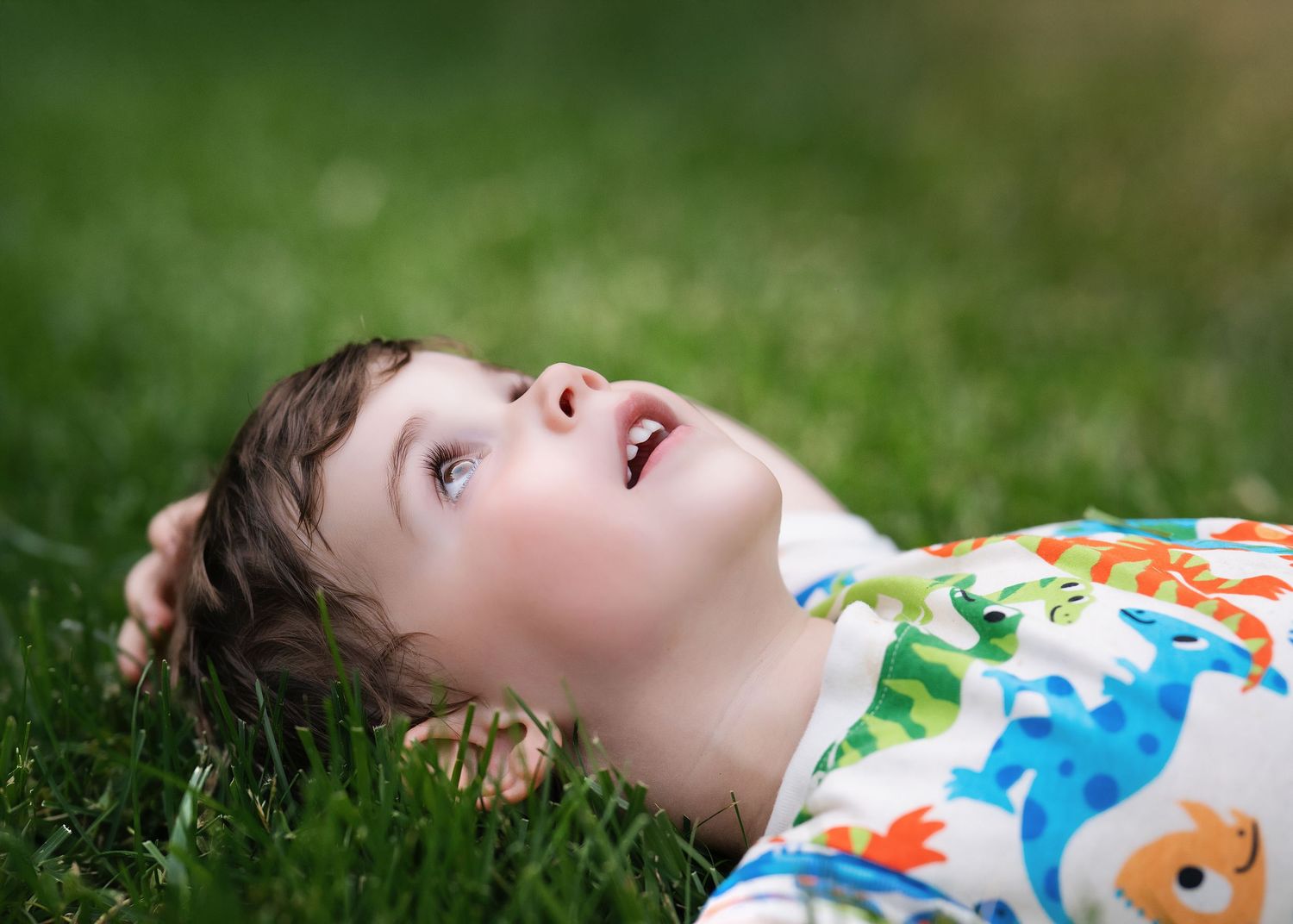 Young child lying on green grass, looking up with a smile, wearing a colorful dinosaur shirt.