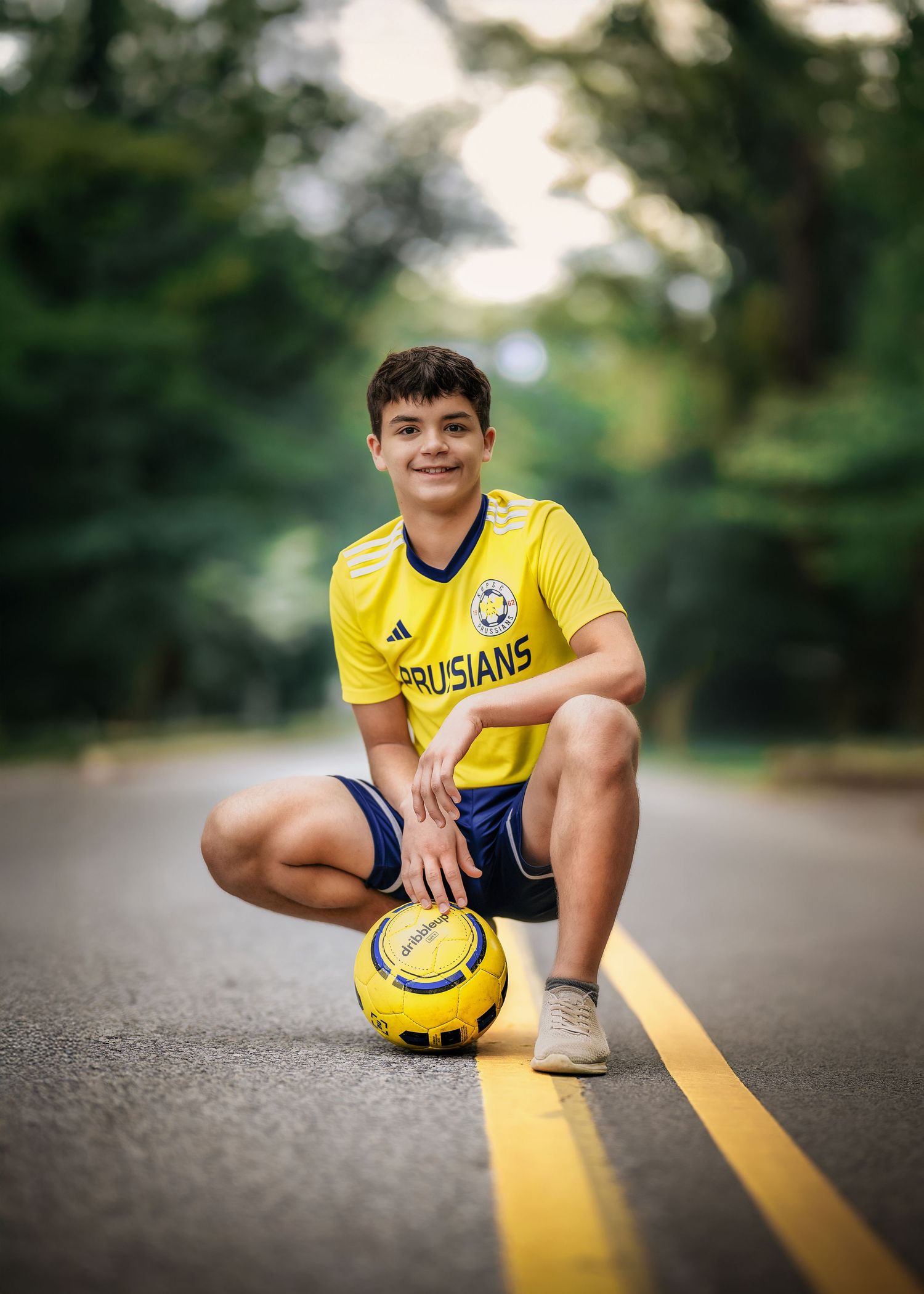 Young soccer player in yellow Prussians jersey crouching on road with ball, surrounded by green trees.