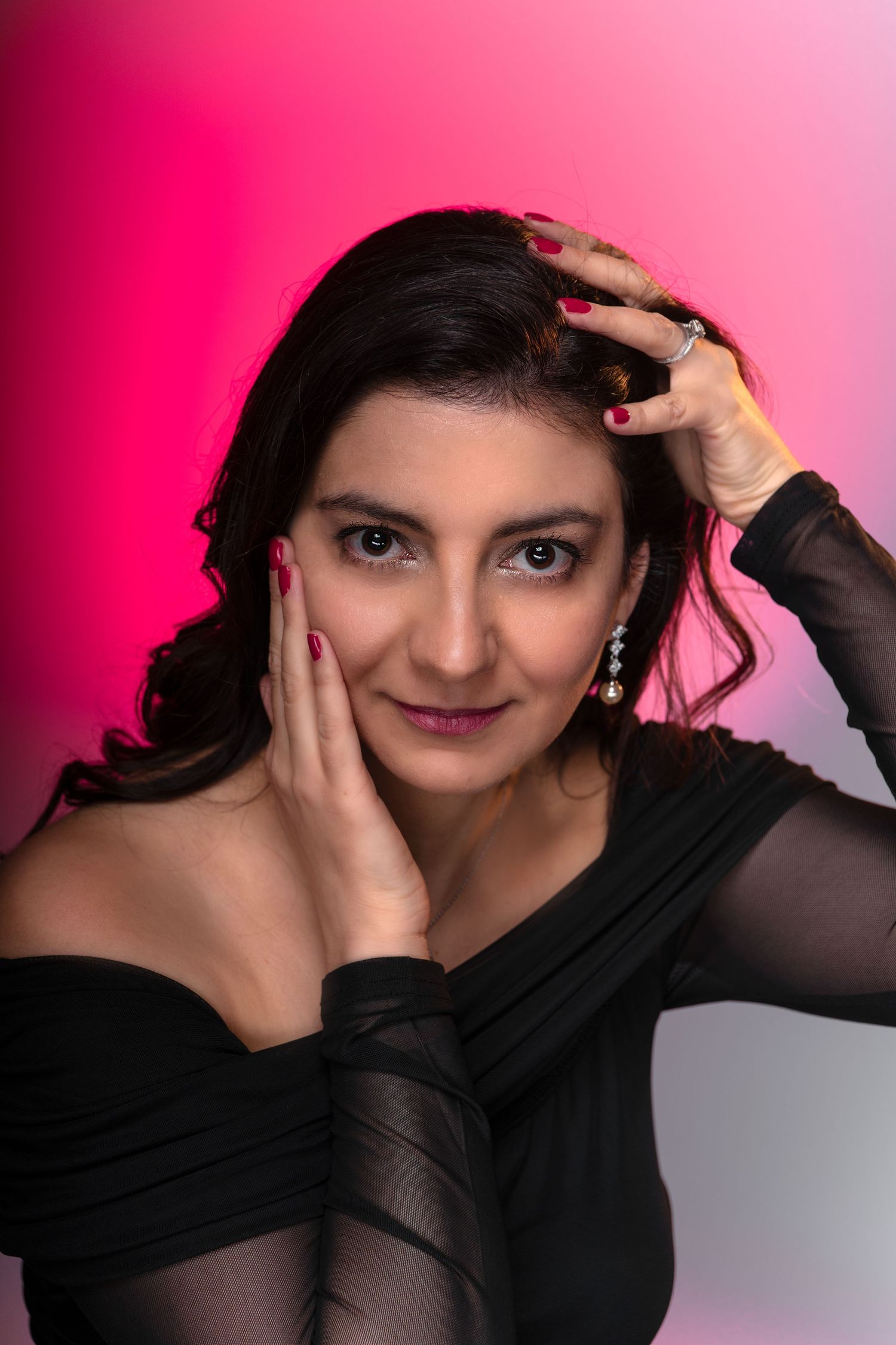 A woman in a black off-shoulder top poses against a pink backdrop, hands framing her face, wearing red nails and drop earrings.
