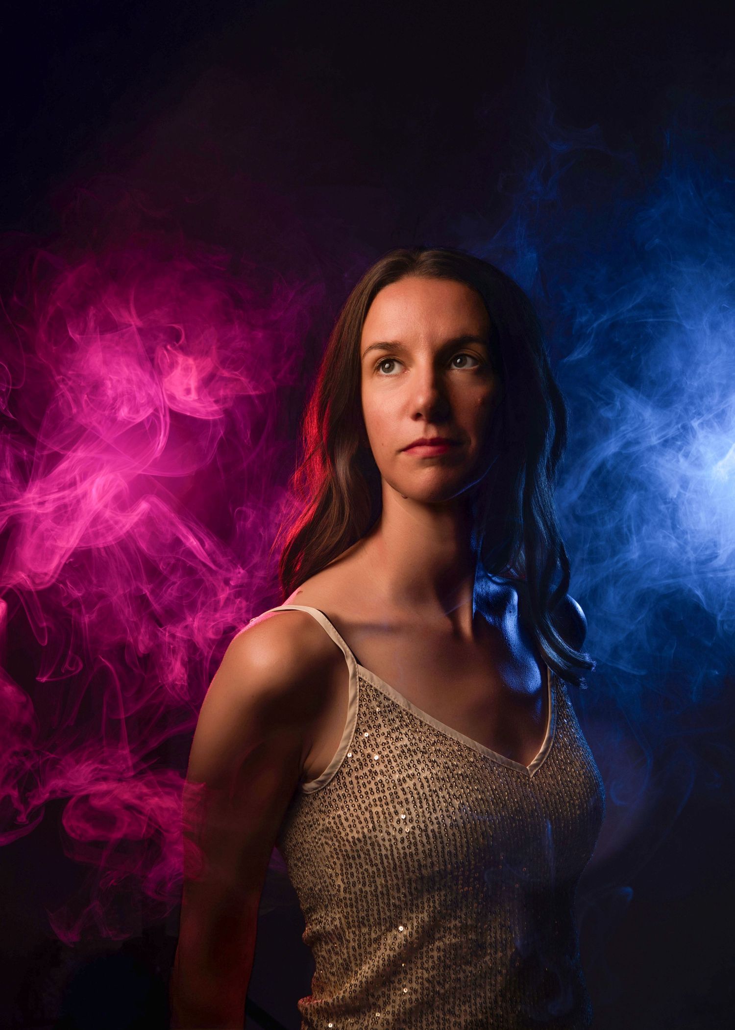 Woman in gold top poses dramatically with vibrant pink and blue smoke in a dramatic women's studio portrait.