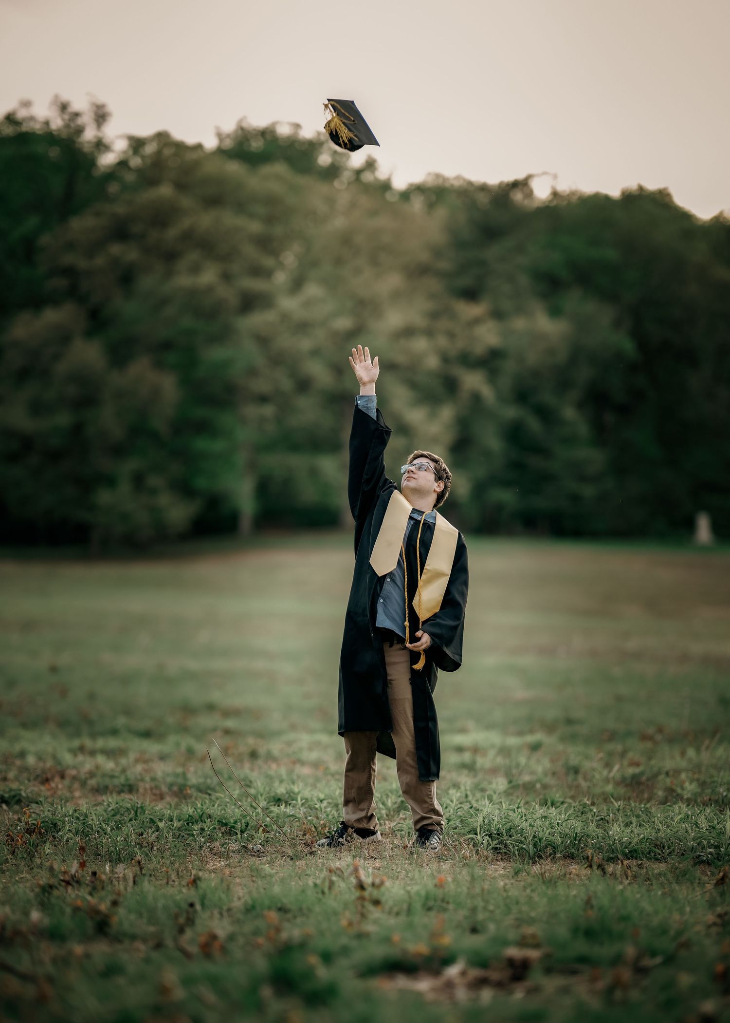 A young graduate in black gown tosses cap in air outdoors in a green field with trees in background.