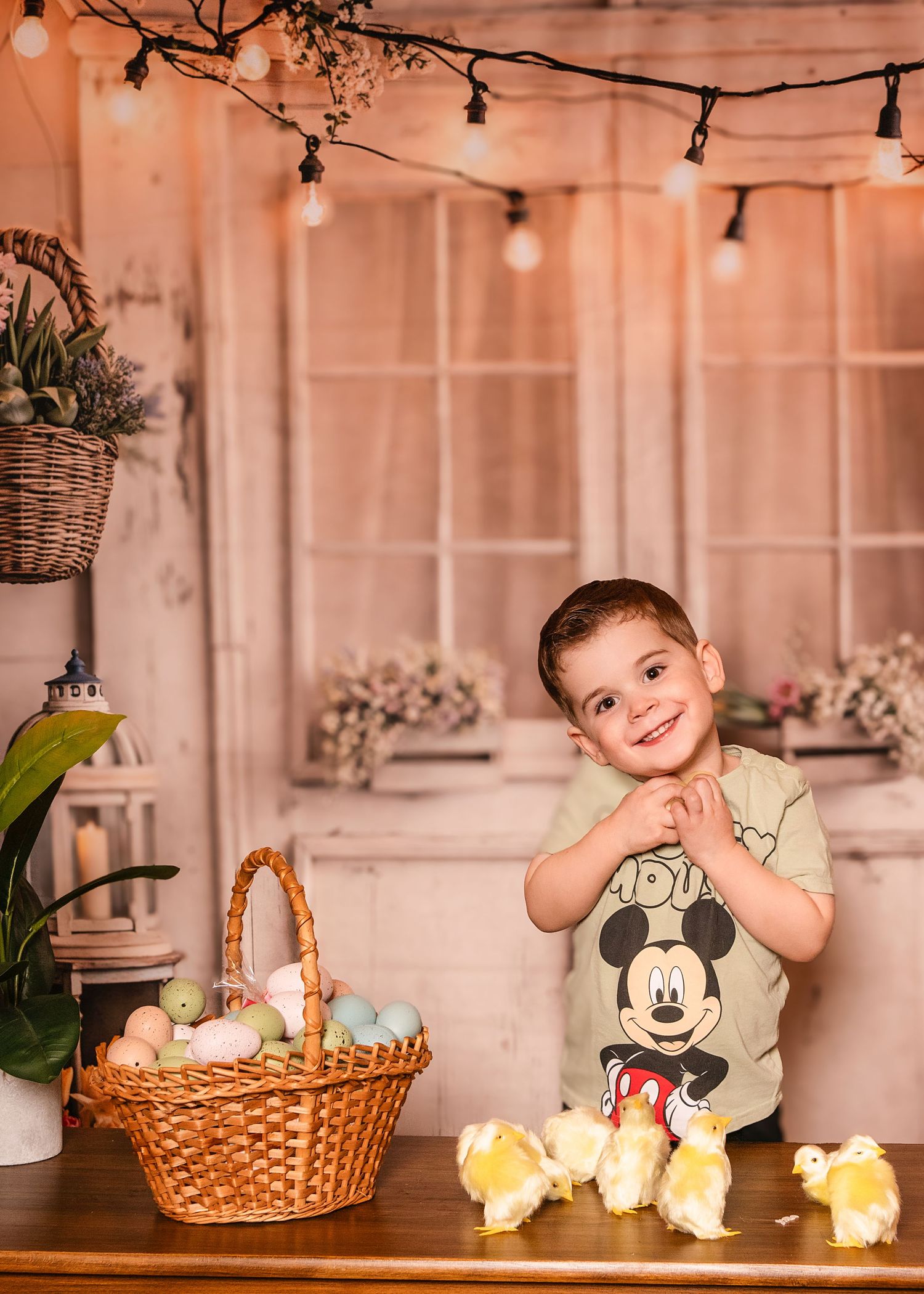 Smiling boy posing next to an Easter basket of colorful eggs and baby chicks on a wooden table during a holiday portrait near Wayne, PA.