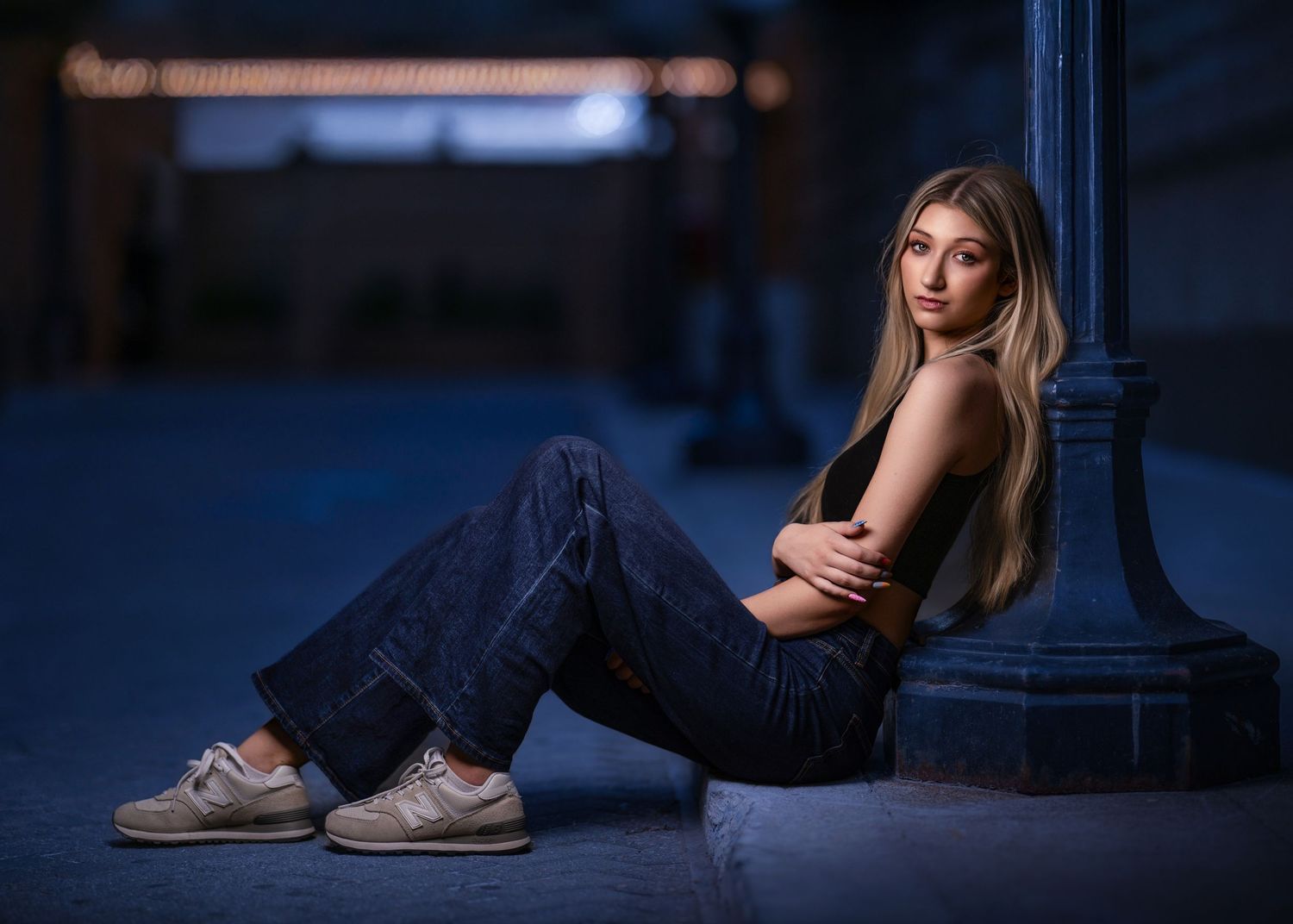 Young blonde woman in jeans and sneakers sitting against a blue pillar outdoors at dusk, arms crossed.