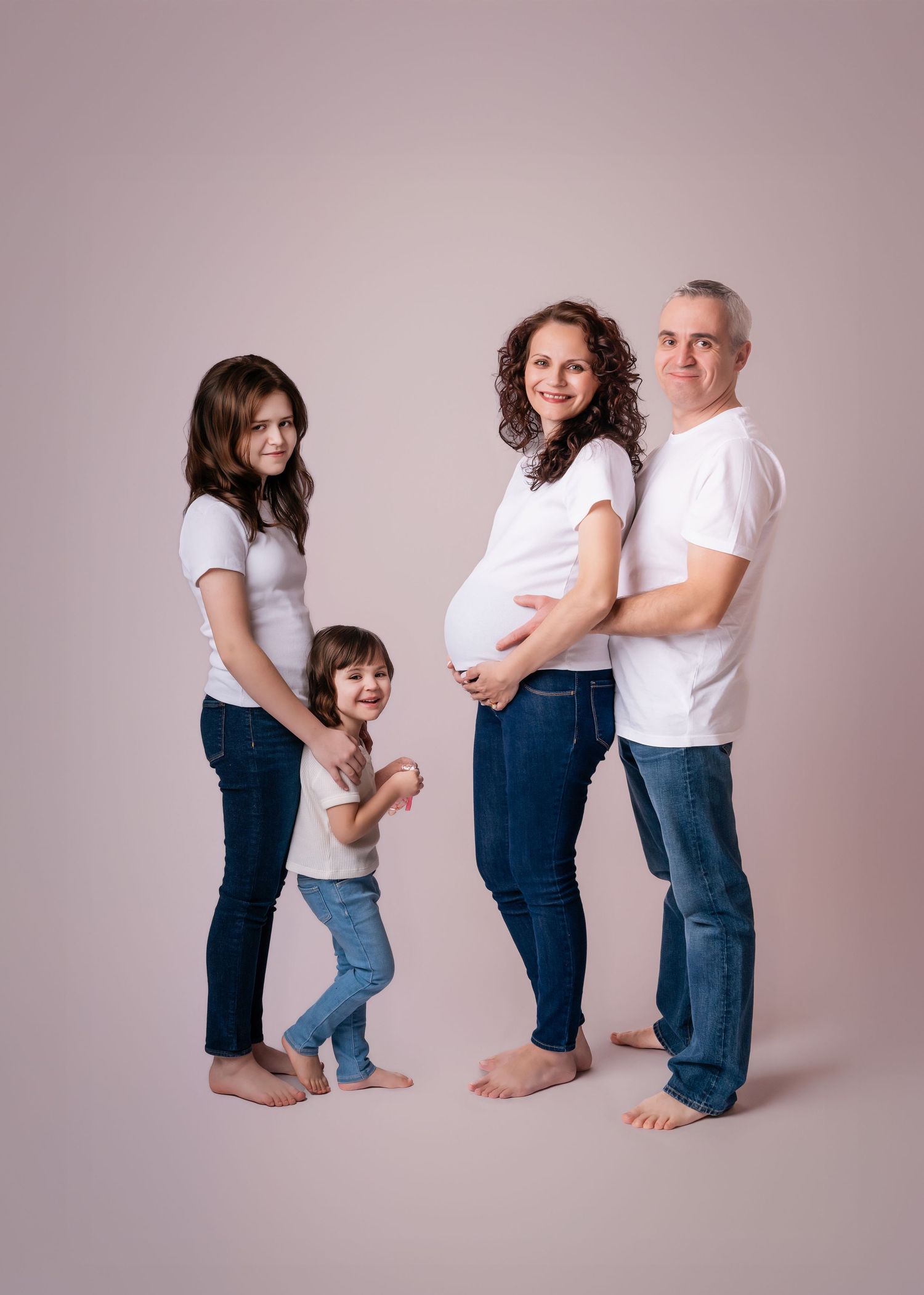 Pregnant woman with family in white shirts and jeans posing for maternity photo on neutral background.