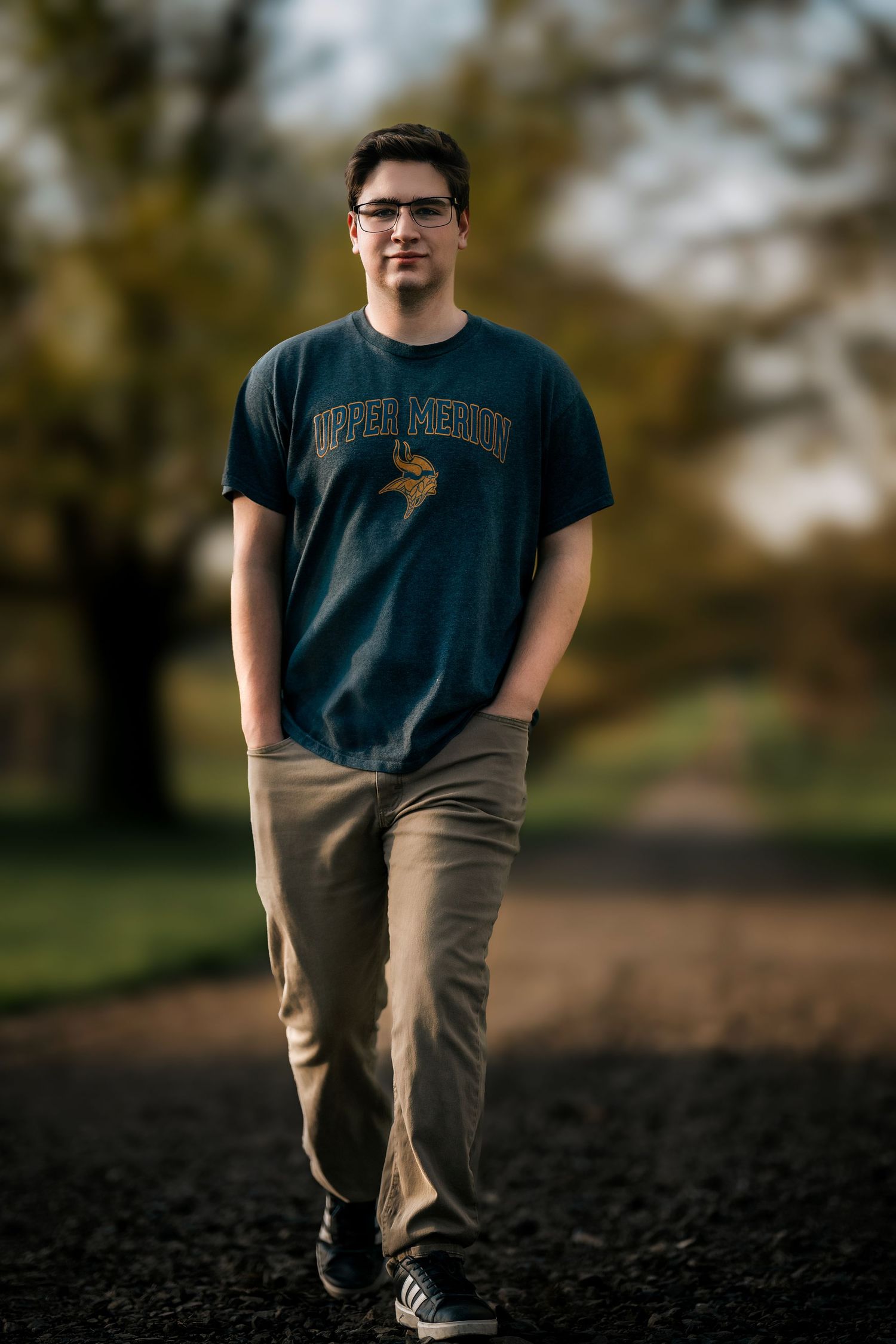 Young man in Upper Merion t-shirt and khakis walks on gravel path in autumn park setting.