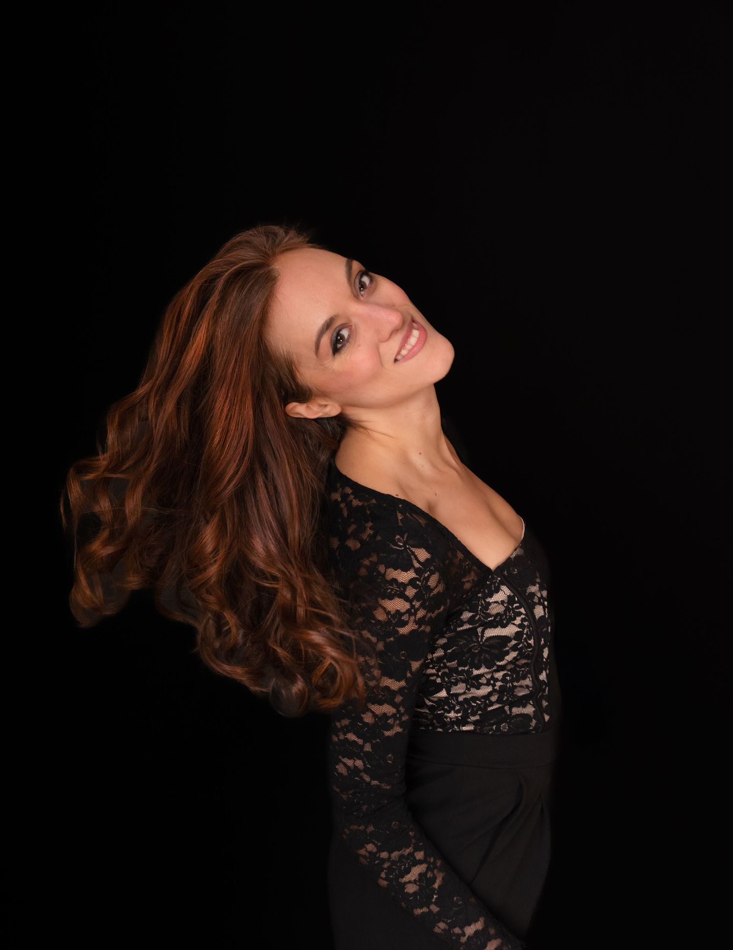 Woman with long auburn hair in black lace dress poses against dark background, hair flowing dramatically.