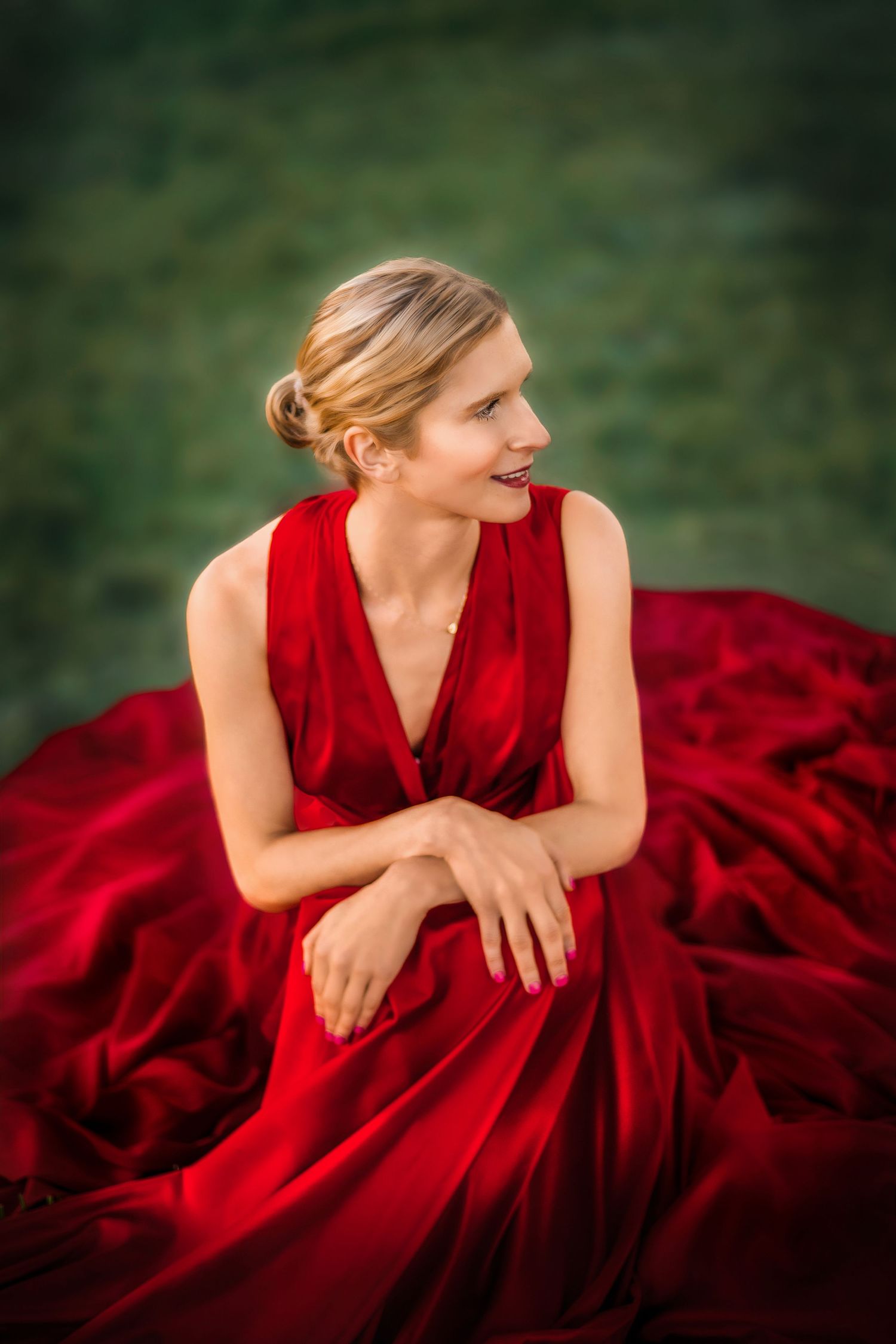 Elegant woman in a flowing red satin gown sitting gracefully on green grass, smiling softly.