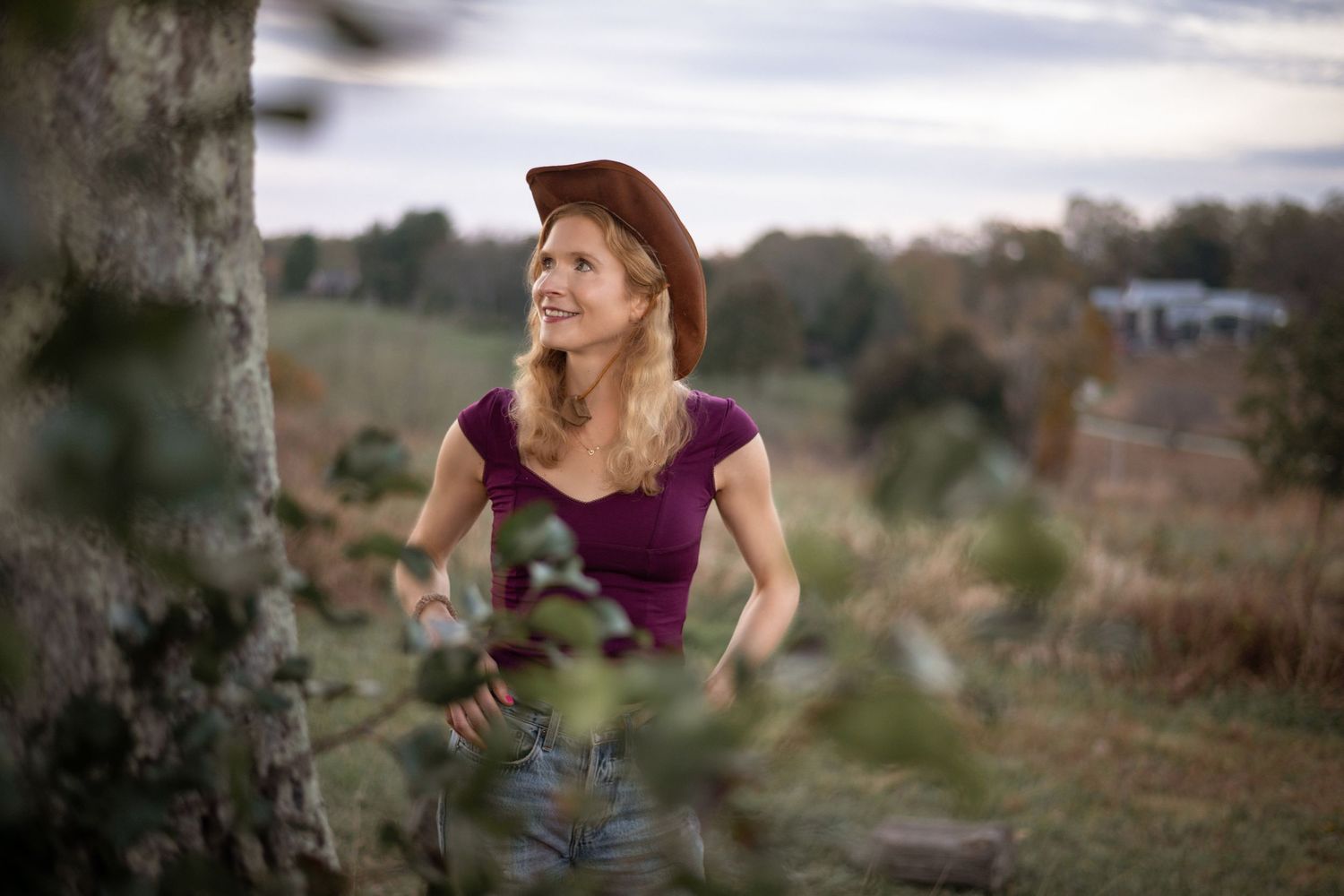Woman in purple top and jeans standing in a golden field at dusk.