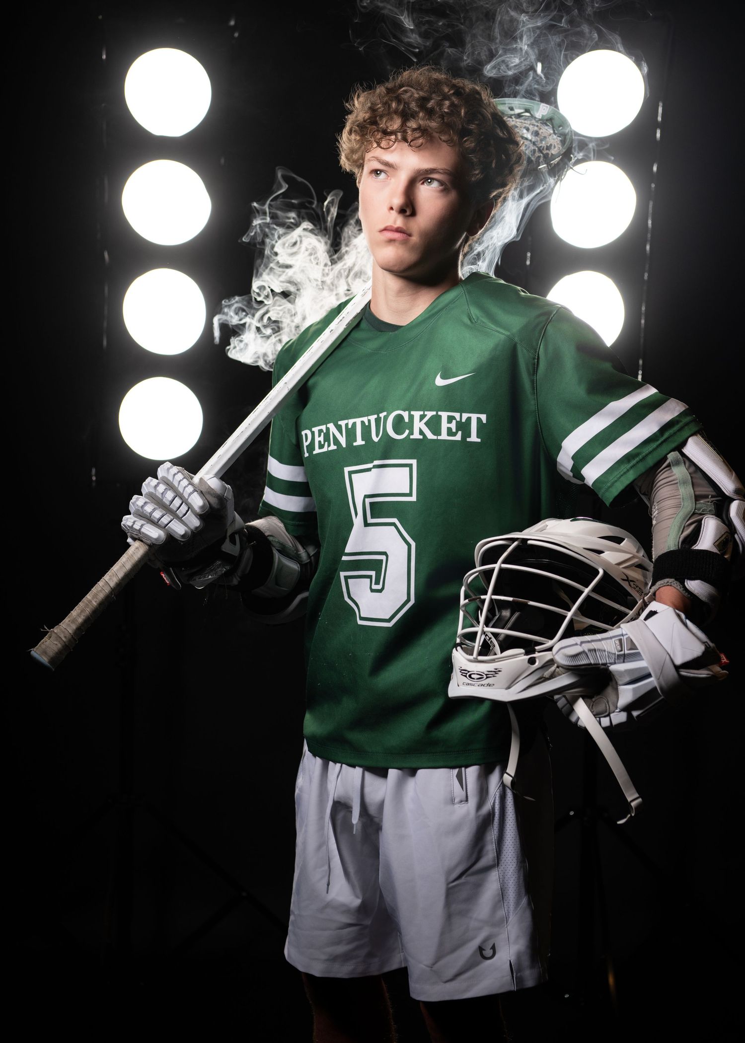 Pentucket #5 lacrosse player in green Nike jersey holding stick and helmet against dramatic studio lighting.