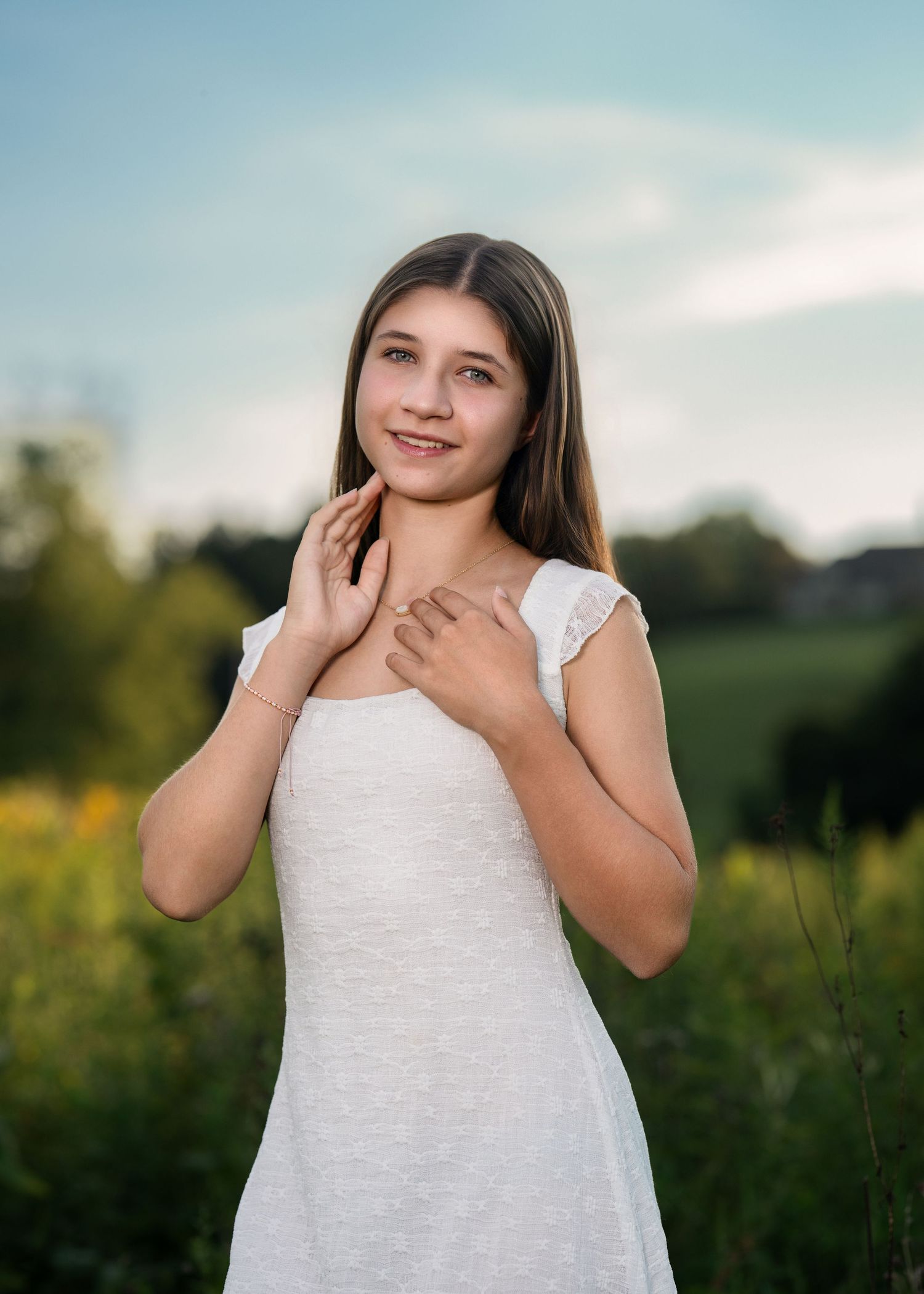 Young girl in white dress posing in a sunny field with yellow flowers and green trees in the background.