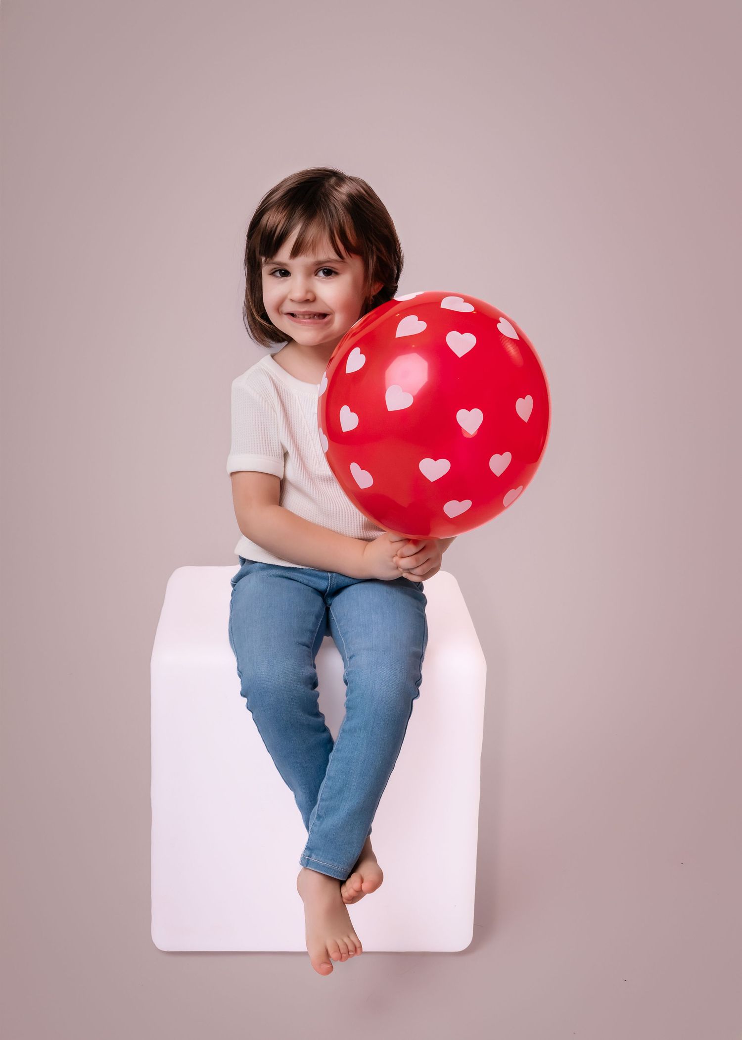 A smiling young girl sits on a white cube holding a red balloon with white hearts on a beige background.
