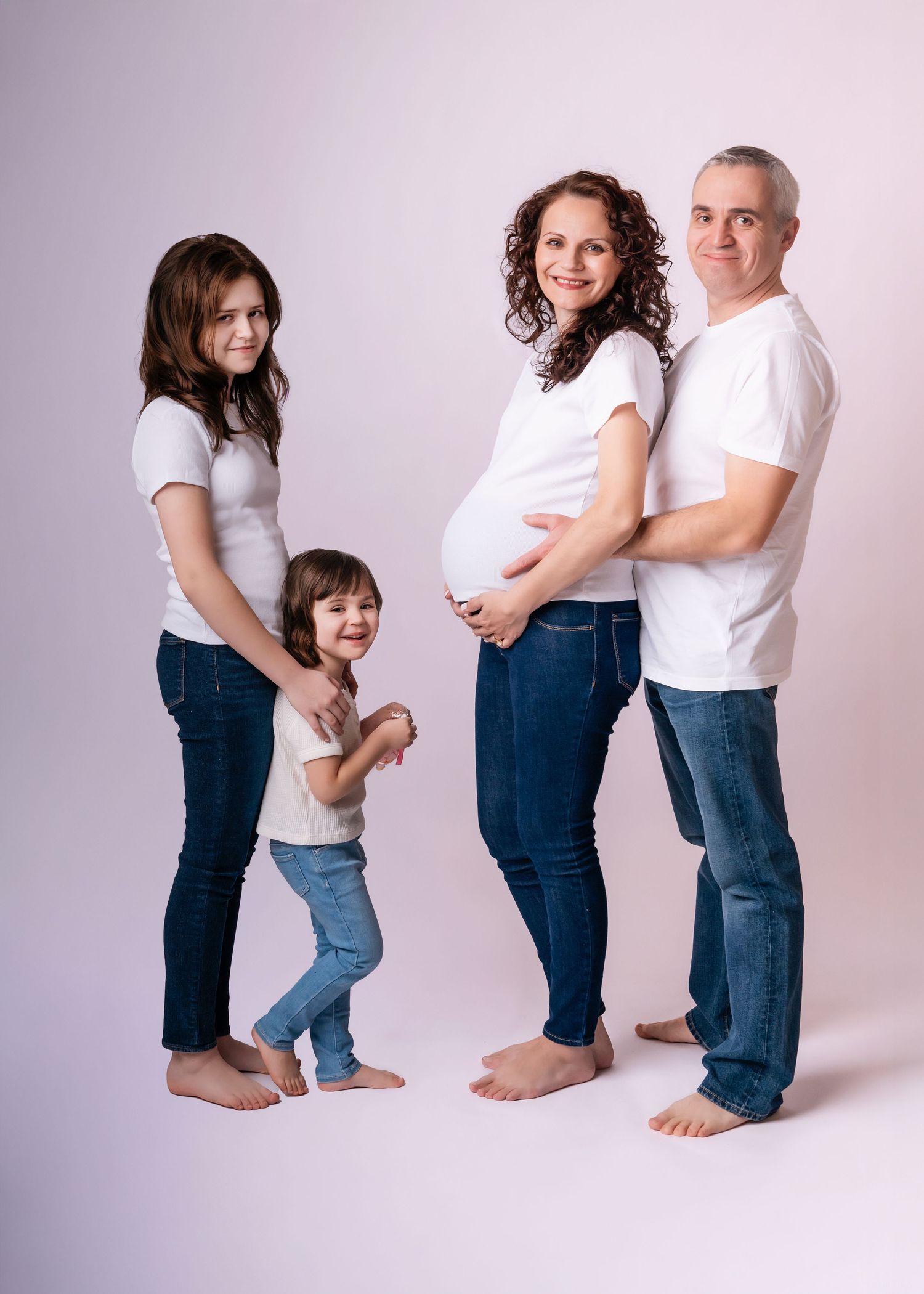 Pregnant woman with family in white shirts and jeans posing for maternity photo shoot in studio.